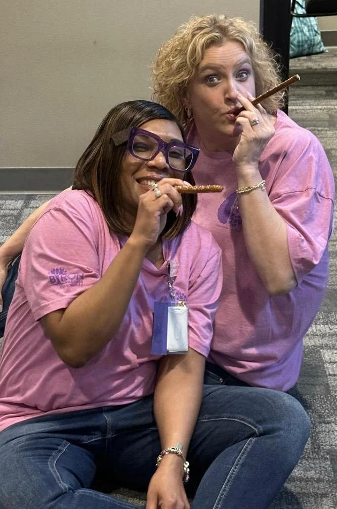 Two women sitting on a carpeted floor, both holding snacks resembling breadsticks, and wearing pink shirts. The woman on the left, with glasses and a badge, is smiling with her eyes closed, while the woman on the right with curly hair is making a pla