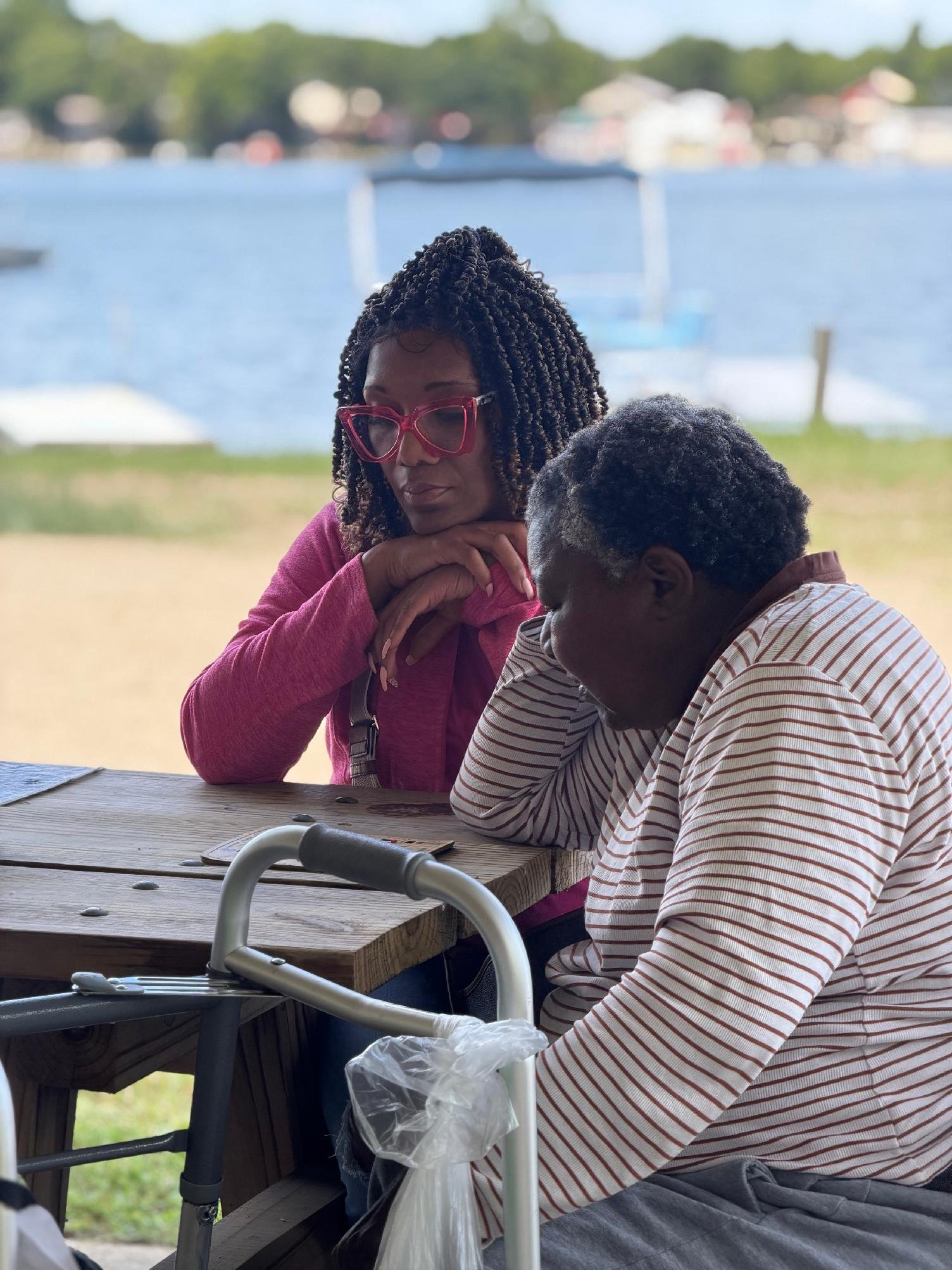 Two women sitting at a picnic table outdoors, one with glasses and the other with short curly hair, having a conversation, with a body of water and boats in the background.