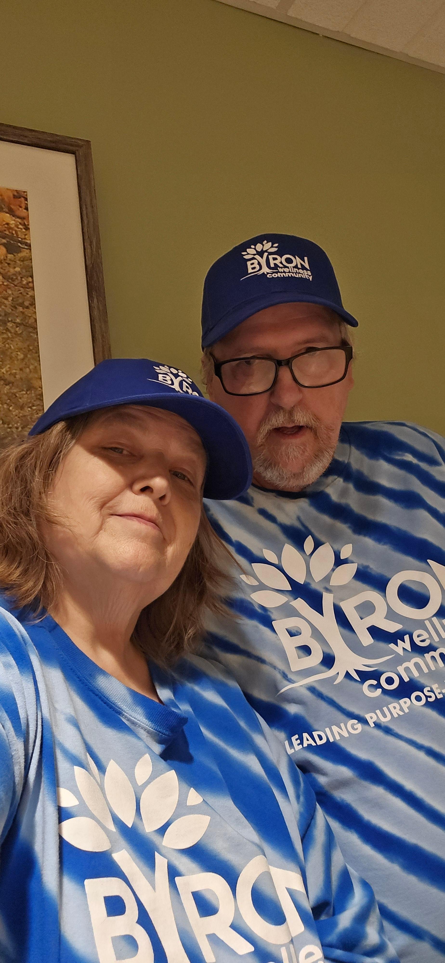 Two people wearing blue tie-dye t-shirts and blue caps with the BIRO logo, taking a selfie indoors against a green wall.