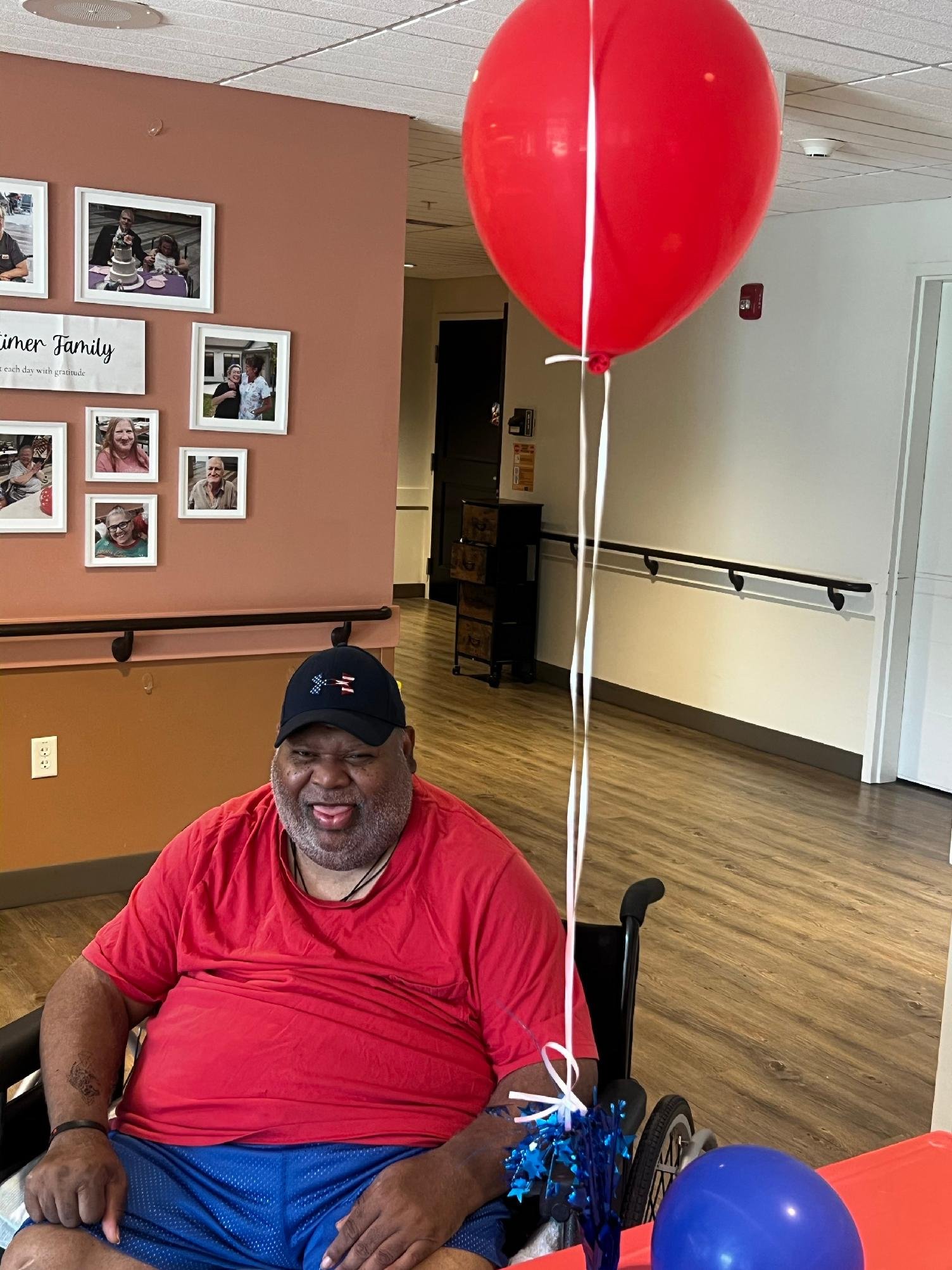 A middle-aged man with a beard is smiling while sitting in a wheelchair, holding blue and white balloons on his lap, with red and blue balloons and a decorated table in front of him, at a celebration event in an indoor hall with wall photos and a sig