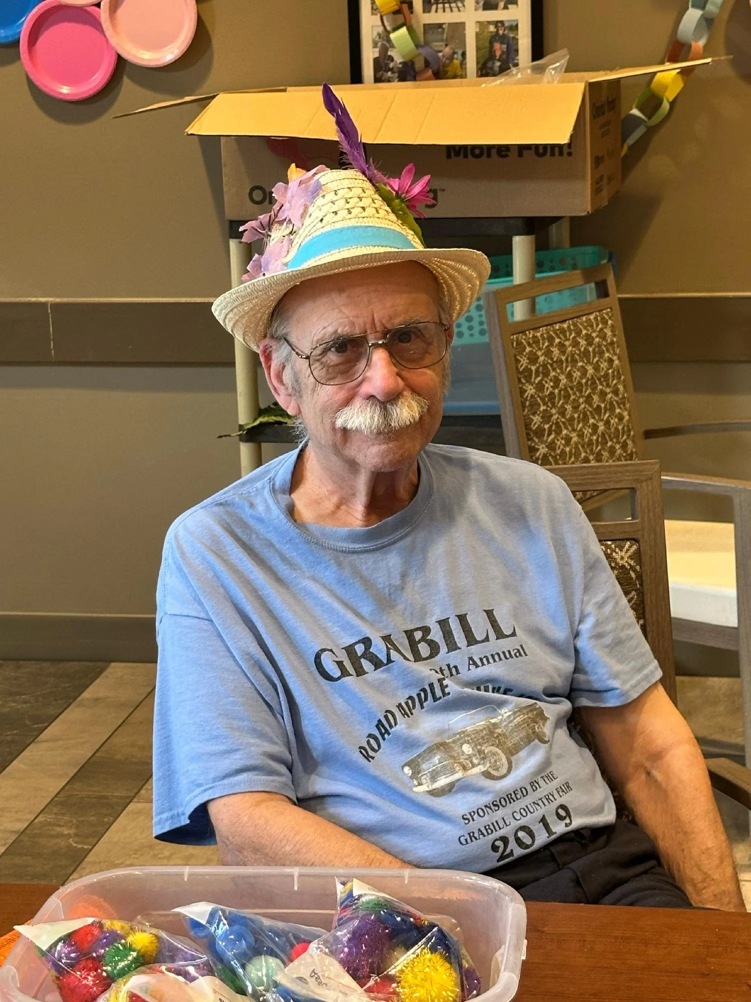 An elderly man wearing glasses and a straw hat decorated with purple and pink feathers, and pink flowers. He is sitting at a table with a plastic container of wrapped candies in front of him, and a cardboard box behind him. The man is wearing a gray 