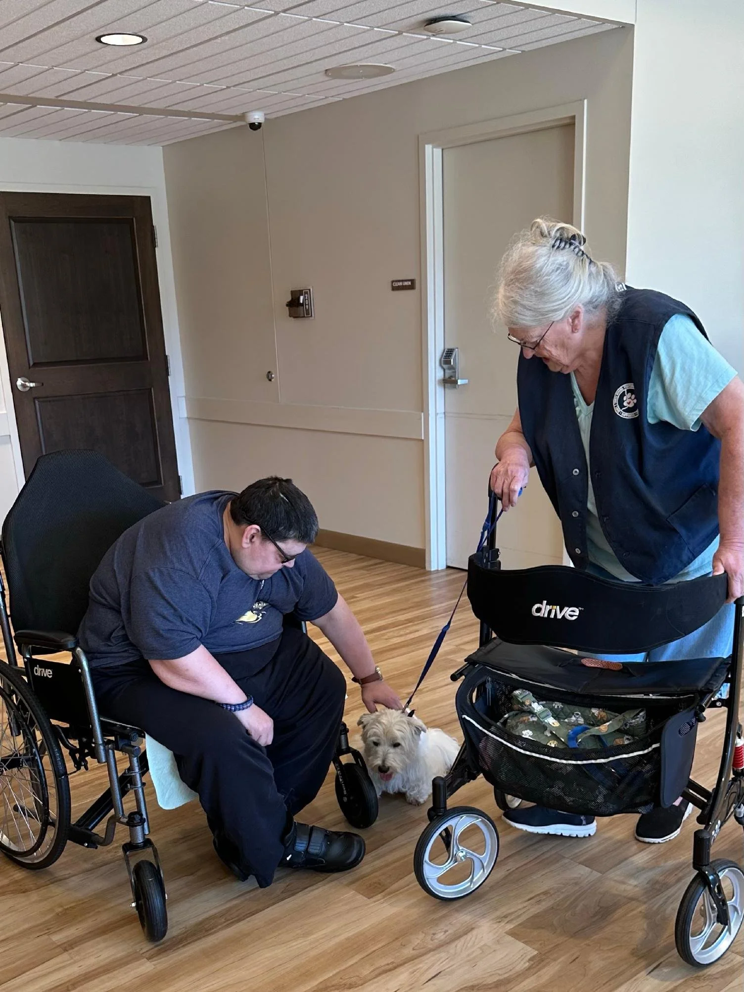 An elderly woman and a young man, both in wheelchairs, are interacting with a small dog in a room with wooden floors and beige walls. The woman is standing and holding a leash attached to the dog, while the young man is reaching toward the dog, who i