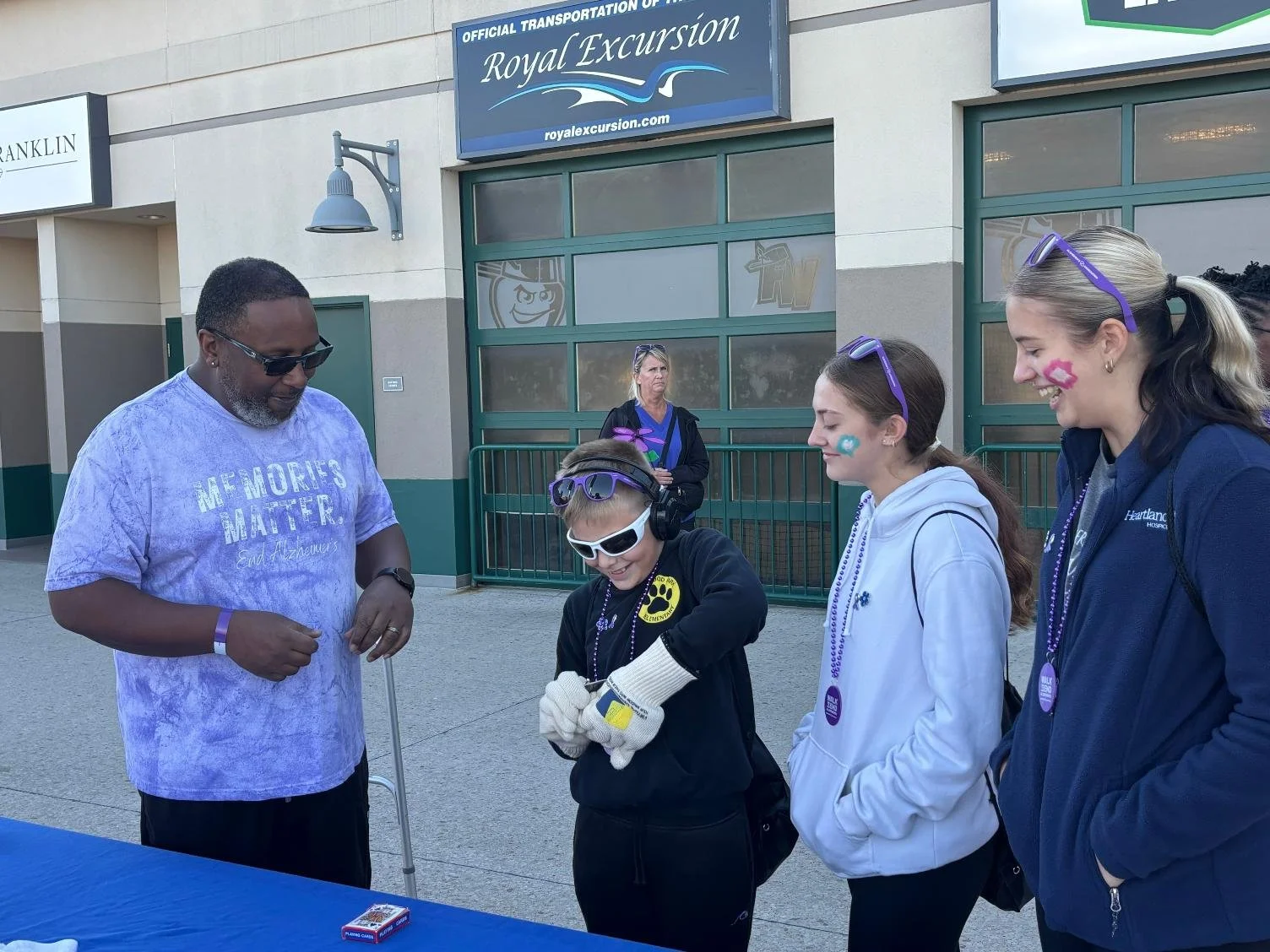 A group of four people standing outside in front of a building with signs. The man on the left is wearing sunglasses, a purple tie-dye t-shirt, and has a cane. The three women are wearing face paint, sunglasses, and dark jackets or hoodies. They are 