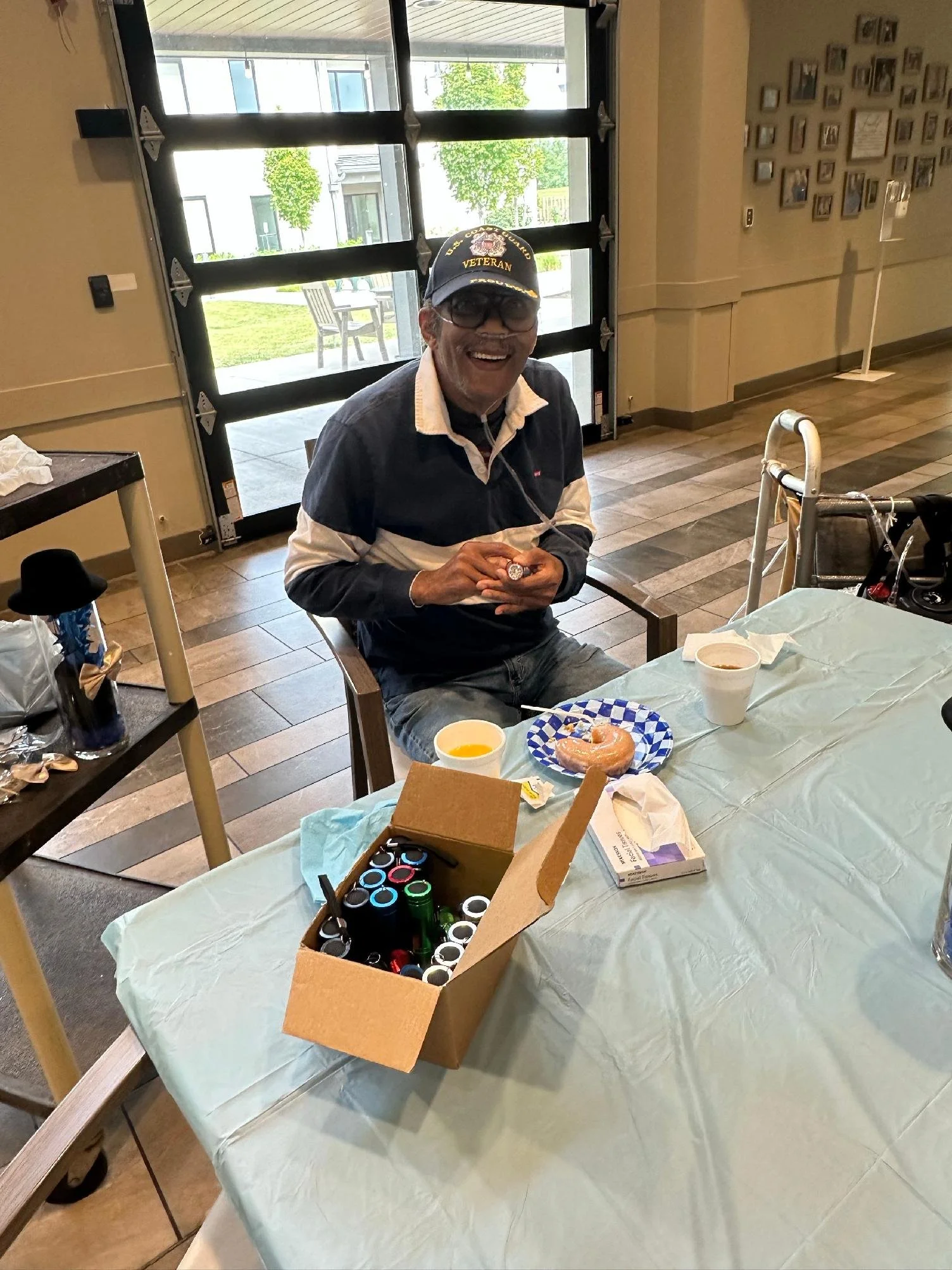 An elderly man with glasses, a veteran cap, and a striped polo shirt is smiling and sitting at a table with donuts and drinks. The table is covered with a light-colored tablecloth, and there is a cardboard box filled with bottled drinks in the foregr