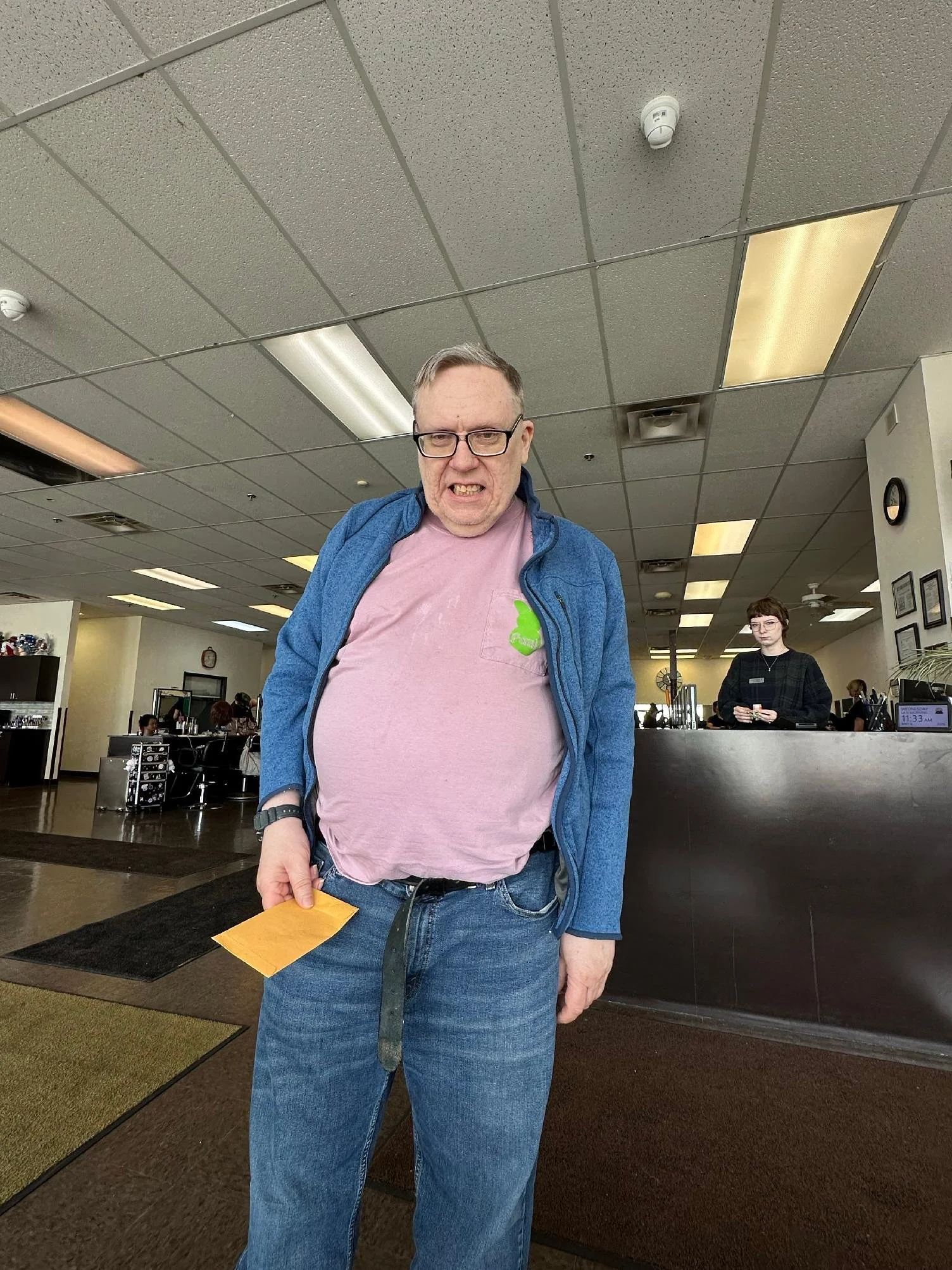 A man with glasses and gray hair, wearing a pink T-shirt and a blue jacket, standing indoors at a counter, holding a yellow envelope, with a woman in black attire in the background, in a space resembling an office or reception area.