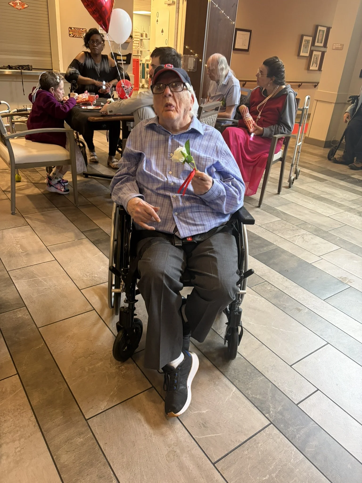 A group of people sitting at a table in what appears to be a restaurant or cafe. There is an elderly man in a wheelchair holding a white flower with red ribbons. Behind him are women and children, some holding balloons and snacks. The setting is warm