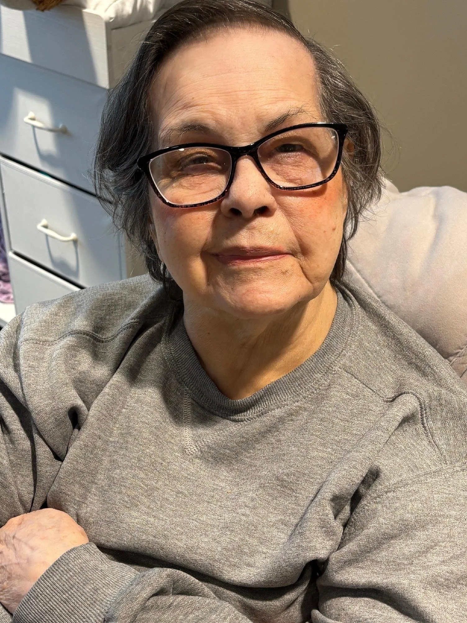 A happy elderly woman with short gray hair and glasses, wearing a gray sweatshirt, sitting on a beige couch near a beige wall with sunlight.