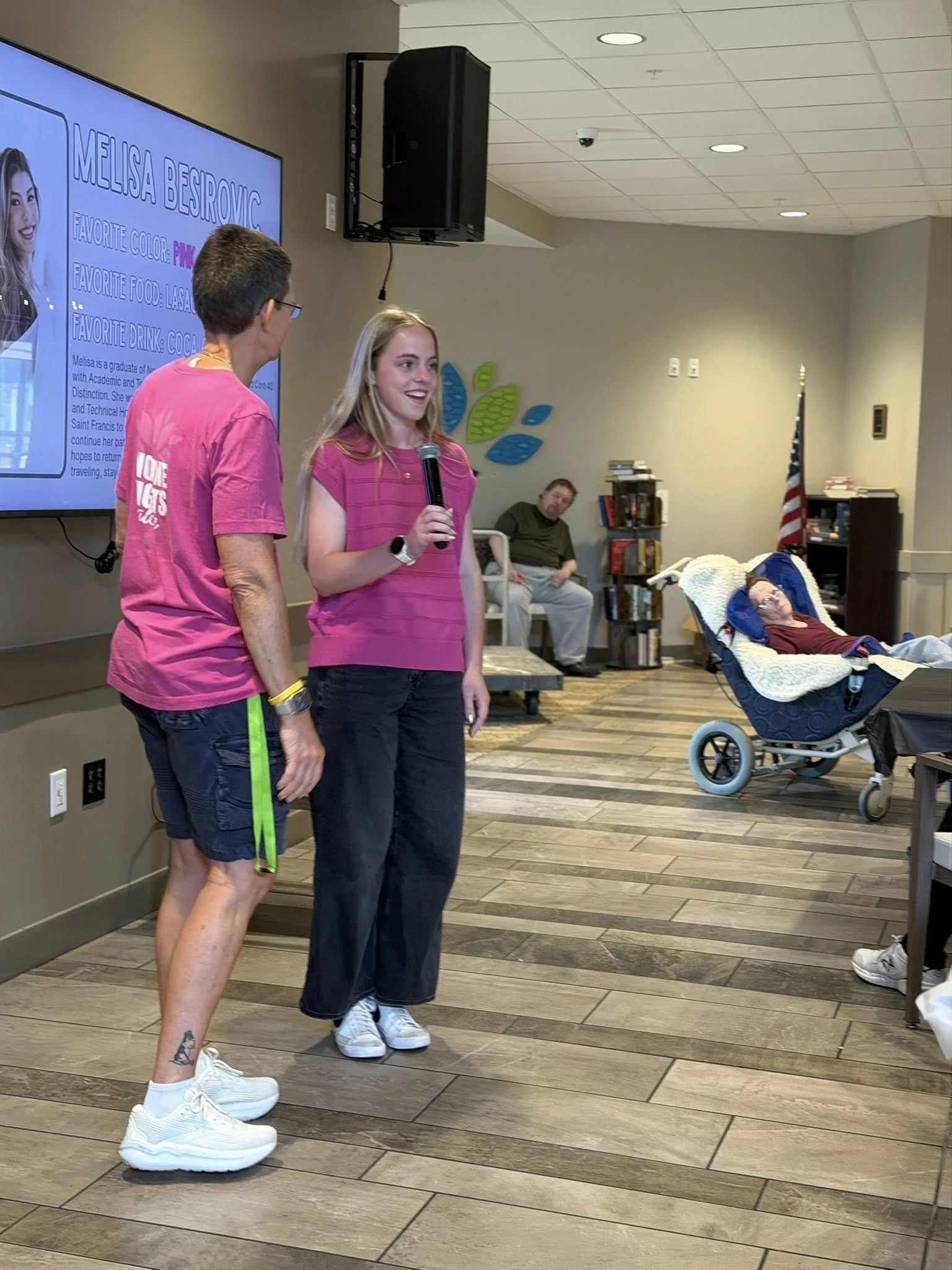 Two women standing and talking in a room with a large TV on the wall. One is holding a microphone, wearing a pink striped shirt and black pants. The other woman, wearing glasses and a pink t-shirt, has a tattoo on her lower leg and is also wearing sn