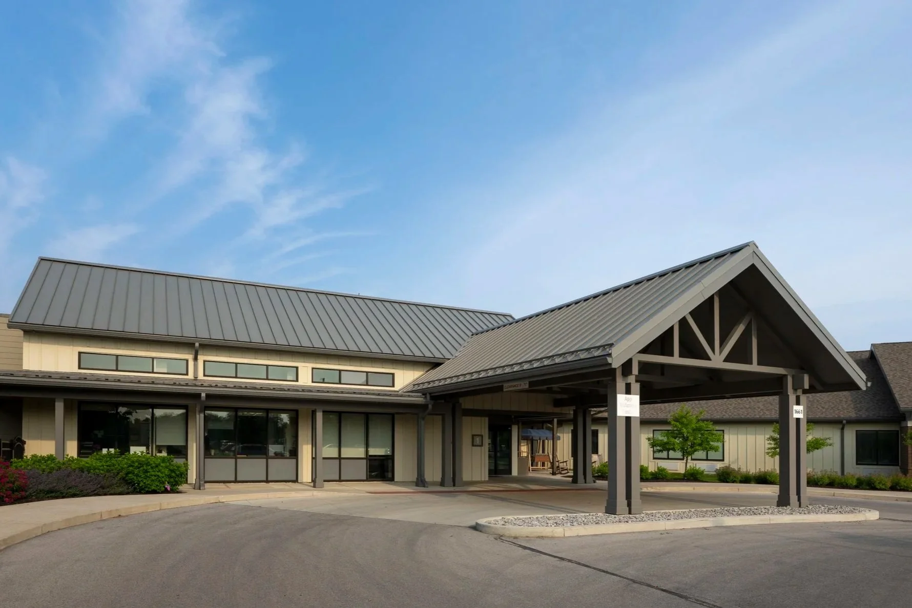 A modern building with a metal roof, large windows, and a covered entrance with supporting columns, surrounded by a landscaped area with small trees and shrubs, under a partly cloudy blue sky.