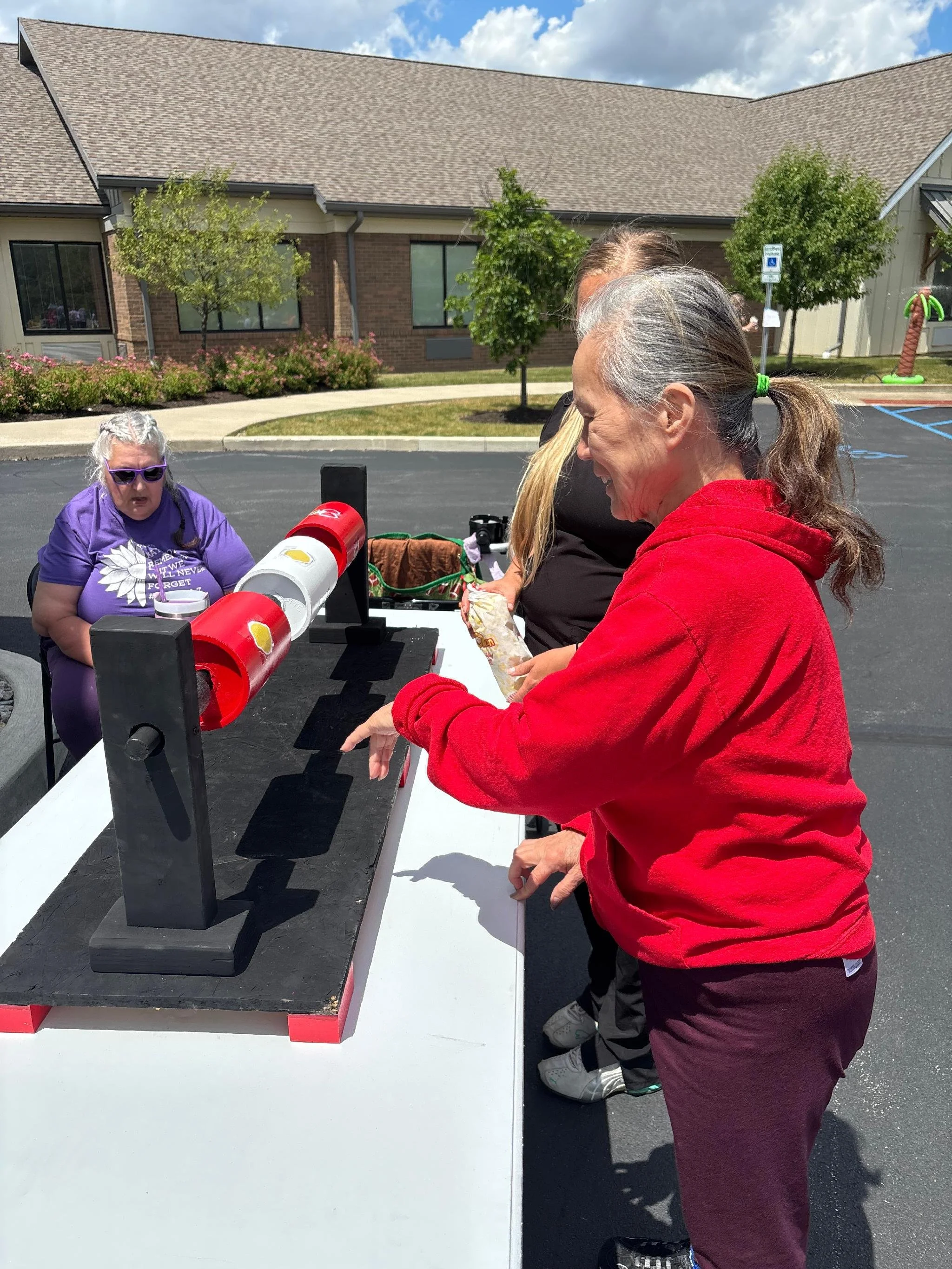 People playing a ring toss game outdoors on a paved surface with a building, trees, and a parking lot in the background.
