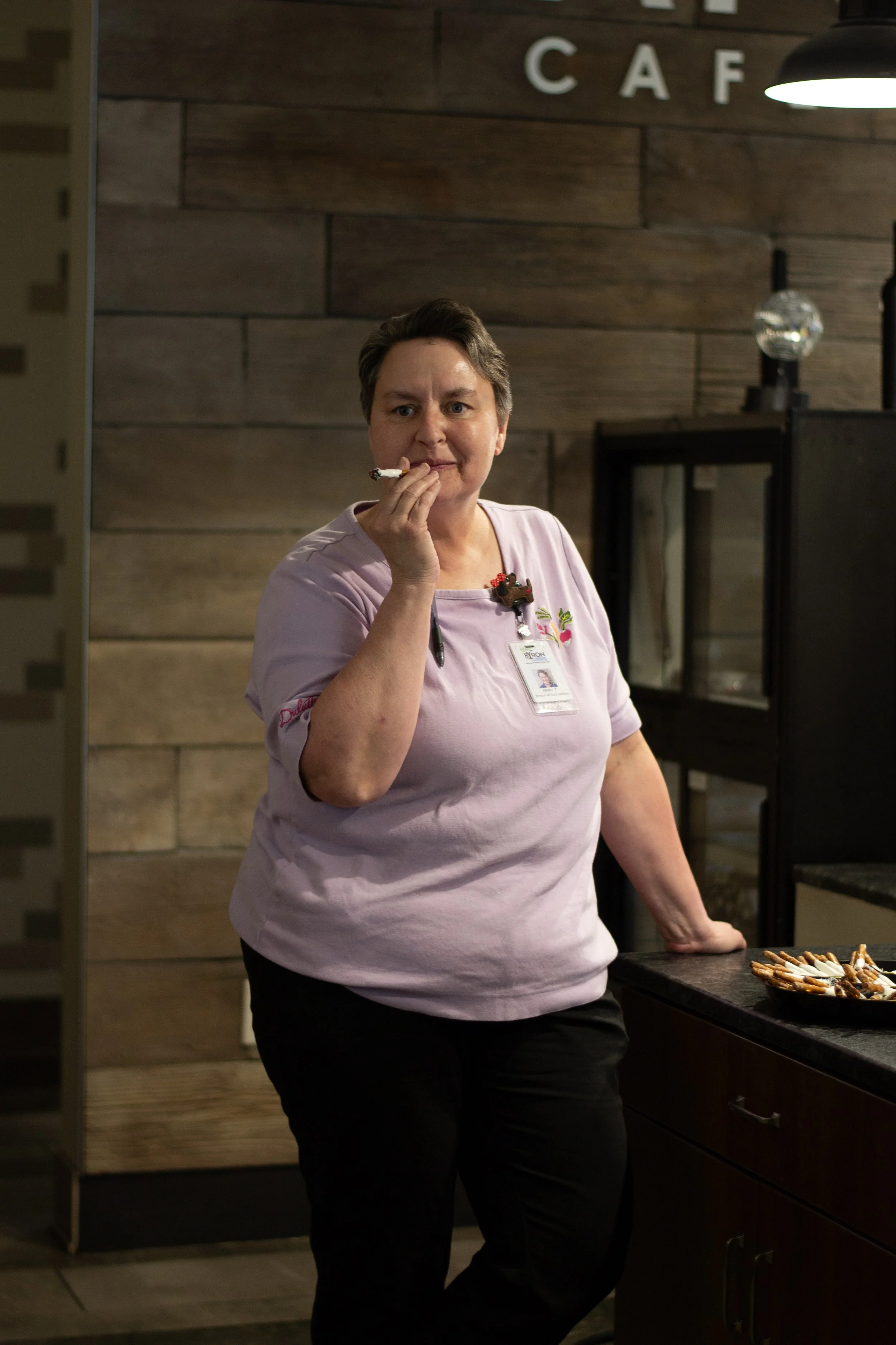 A woman in a pink shirt with a name tag and a small plush bear pin is holding a cigarette, standing near a table with pretzels in a dimly lit café or kitchen with wooden wall decor.