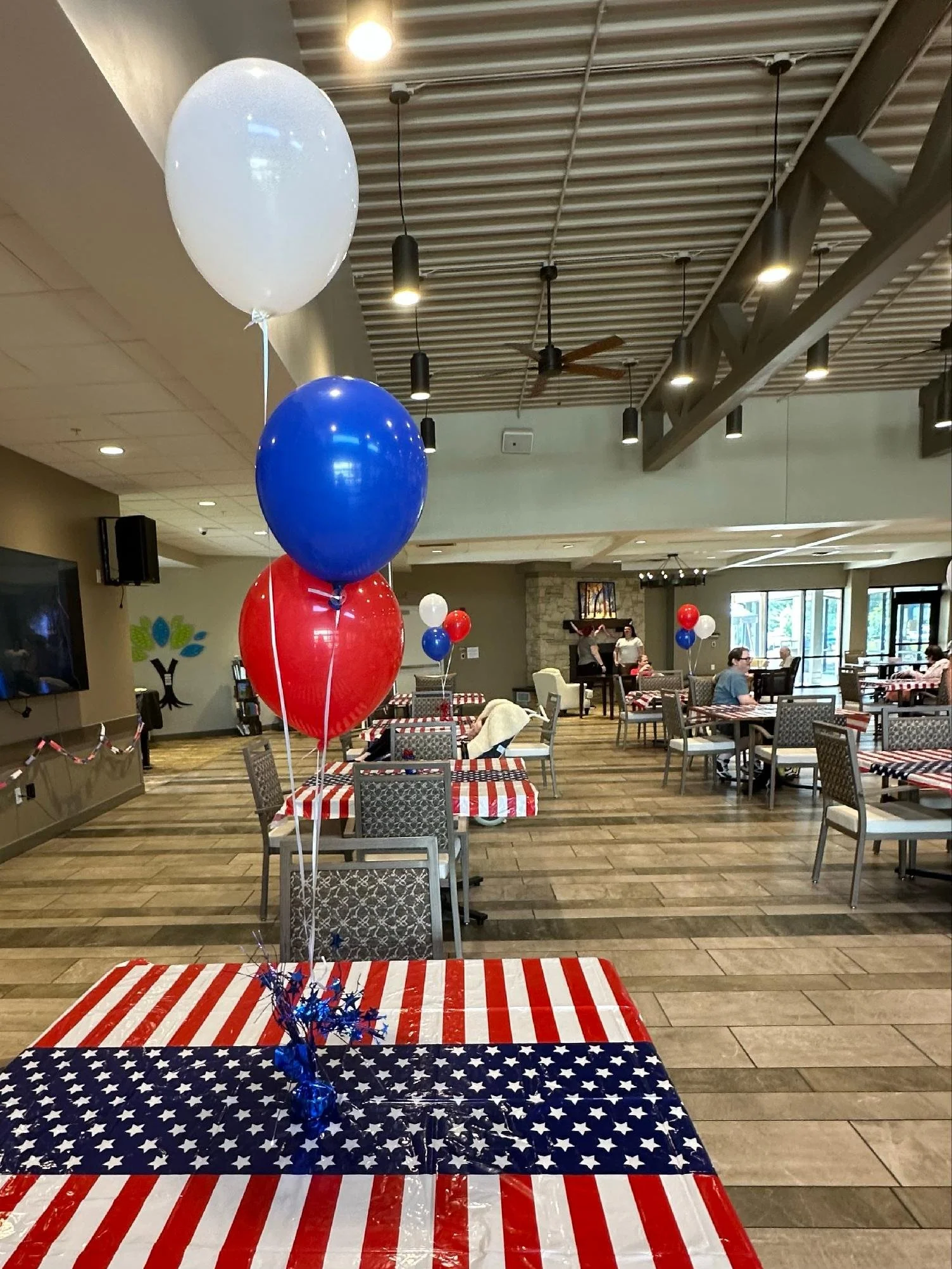 Indoor space decorated with patriotic themed balloons, including white, blue, and red balloons, and tables with red, white, and blue tablecloths featuring stars and stripes, indicating a celebration or event.