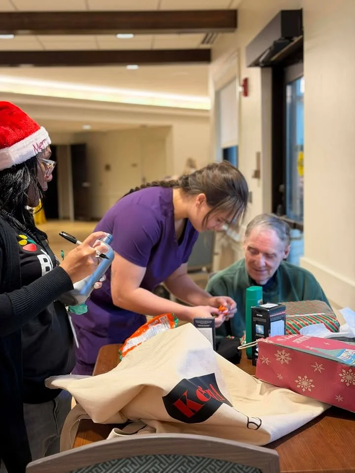 Three people around a table opening Christmas gifts, one wearing a Santa hat, in a well-lit room.