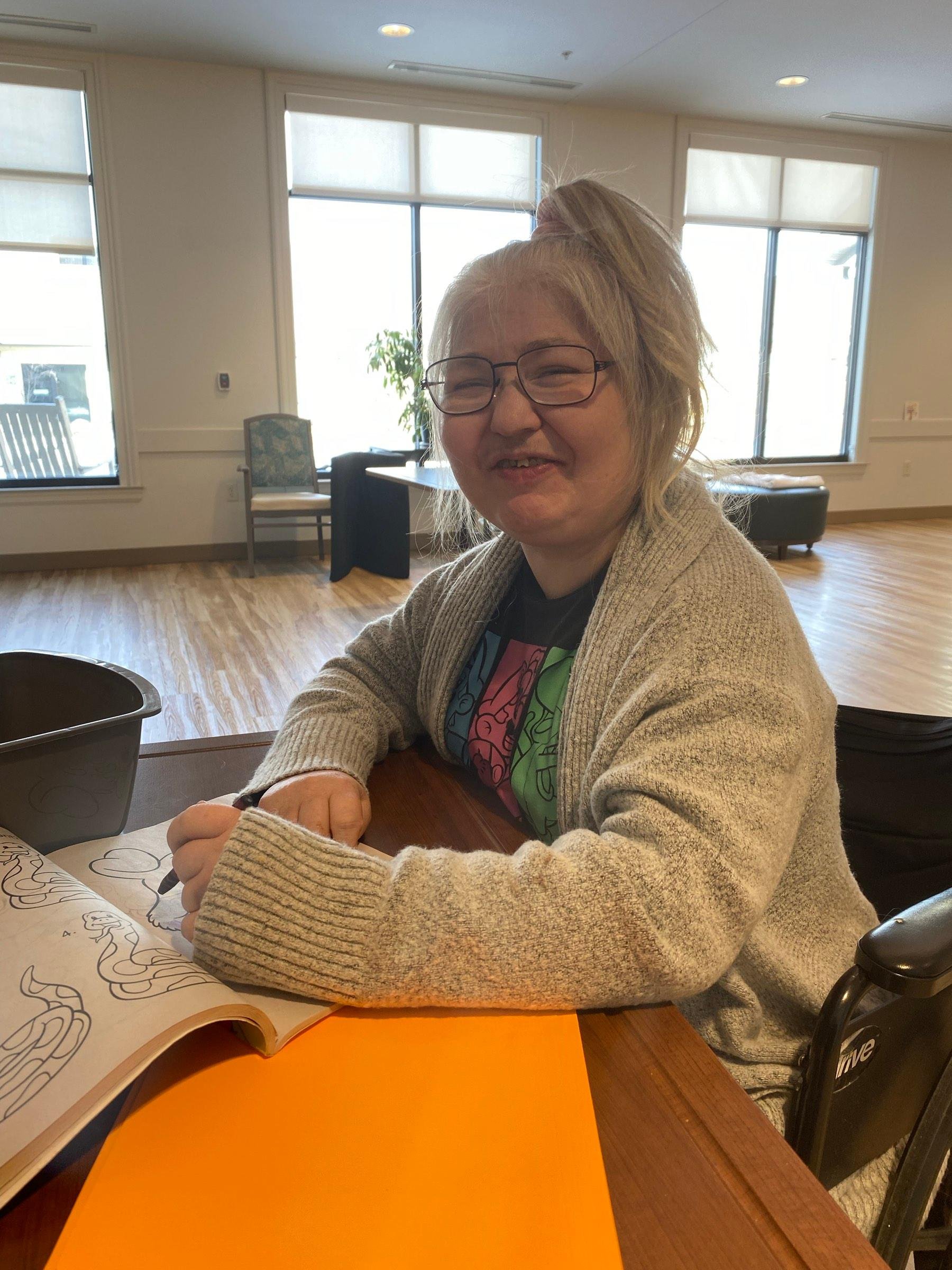 Older woman with glasses and blond hair in a top knot, smiling with eyes closed, seated at a table coloring with a marker in a room with large windows, wooden flooring, and minimal furniture.
