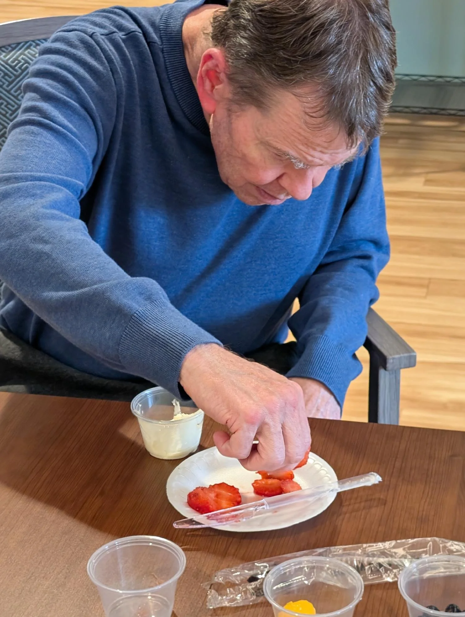 An elderly man in a blue sweater is cutting strawberries on a paper plate at a wooden table. There are small cups with other fruits on the table.
