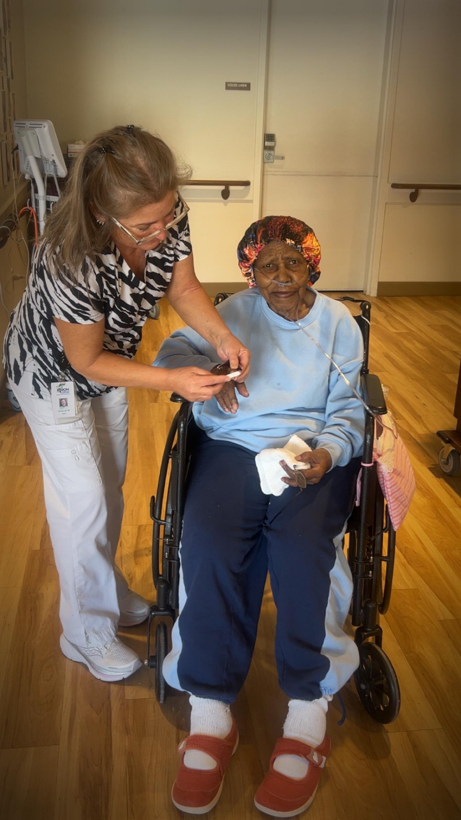 A healthcare worker assisting an elderly woman in a wheelchair. The woman is wearing a colorful hair cap and a nasal oxygen tube. The healthcare worker is holding her hand and appears to be talking to her. The setting is a medical or care facility with wood flooring and closed doors.