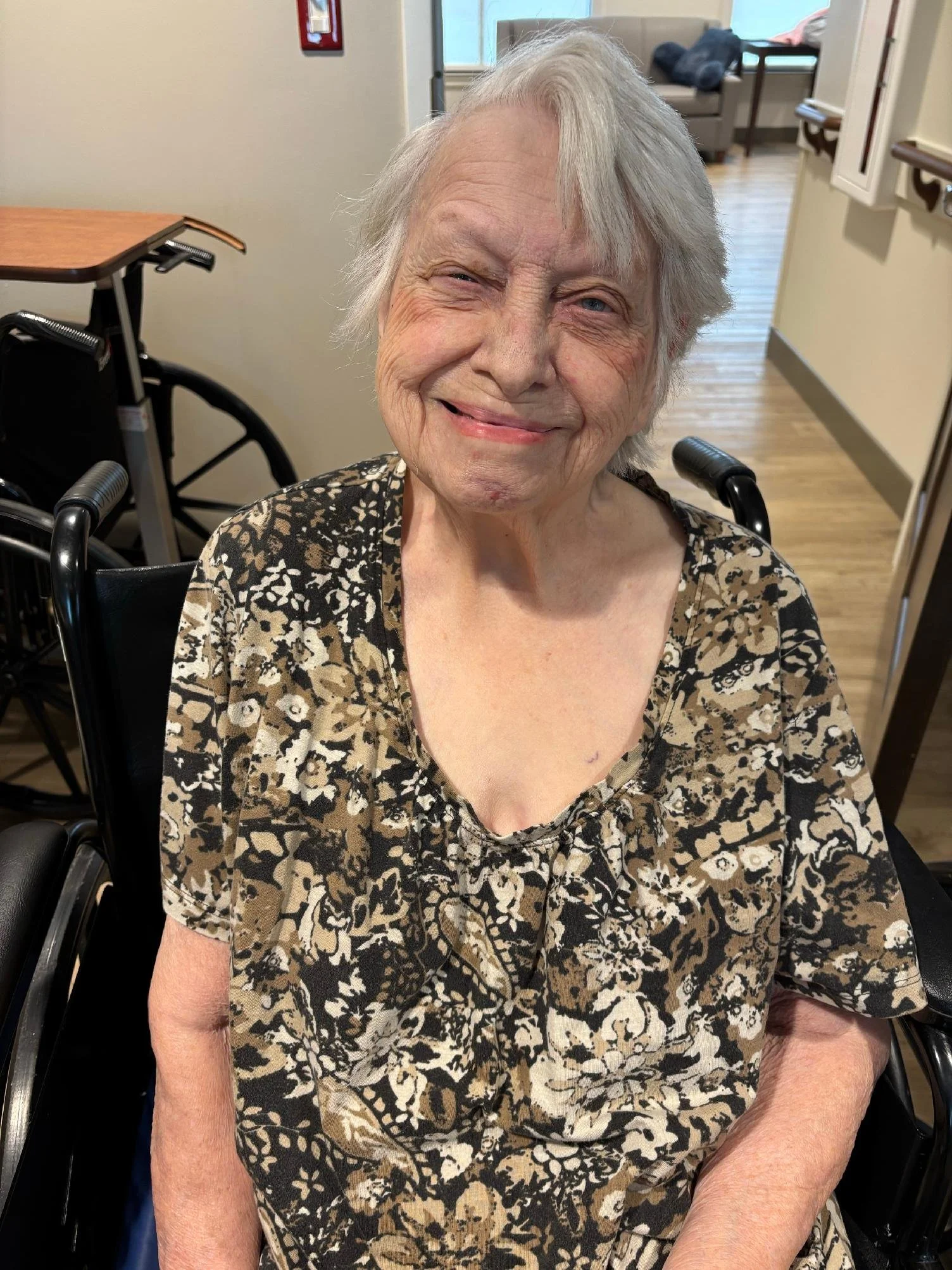 A smiling elderly woman with short white hair sitting in a wheelchair in a nursing home or assisted living facility.