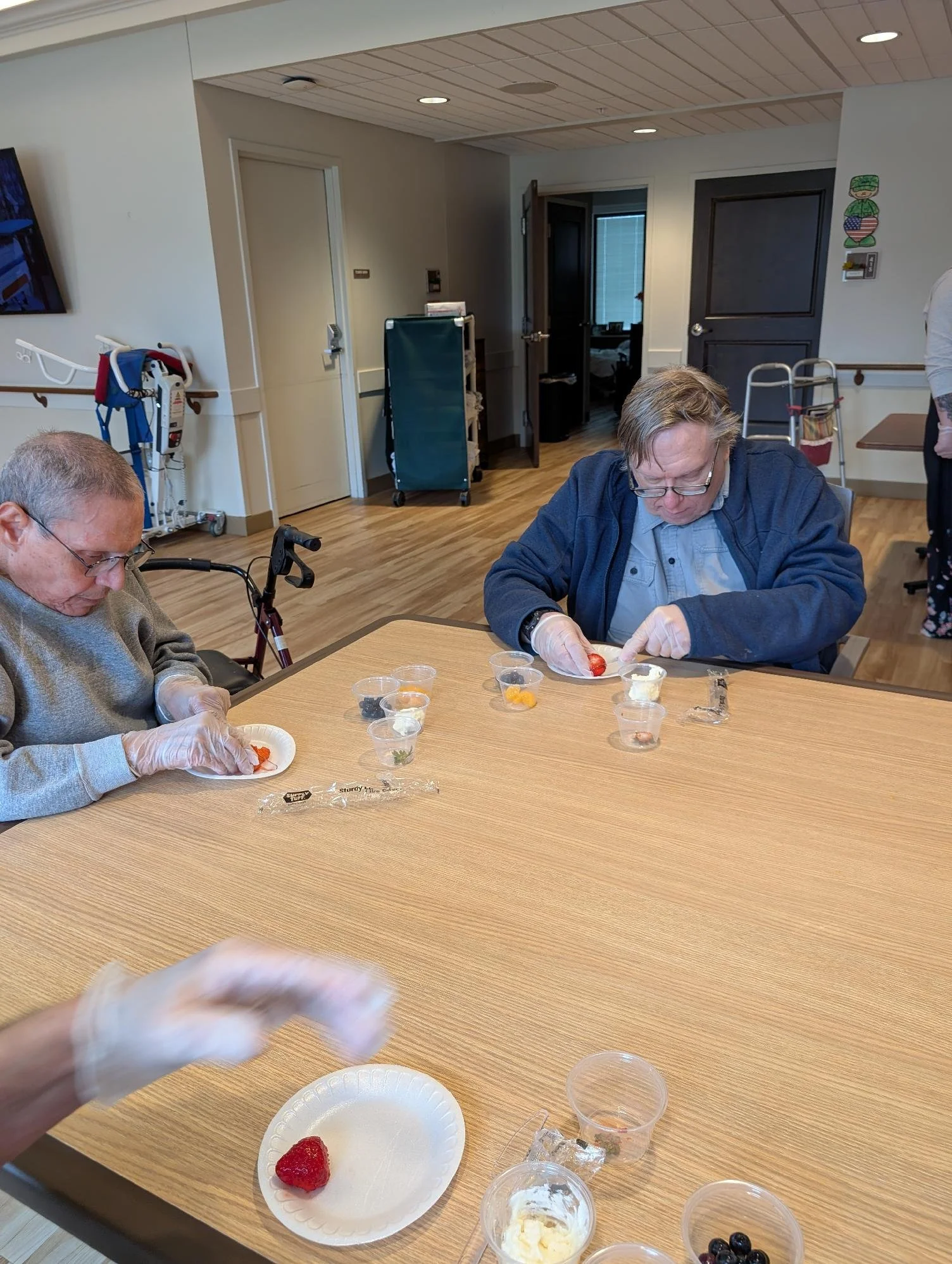 Two elderly men in a room with medical equipment are sitting at a large wooden table, preparing small servings of dessert or snacks from various bowls and cups.