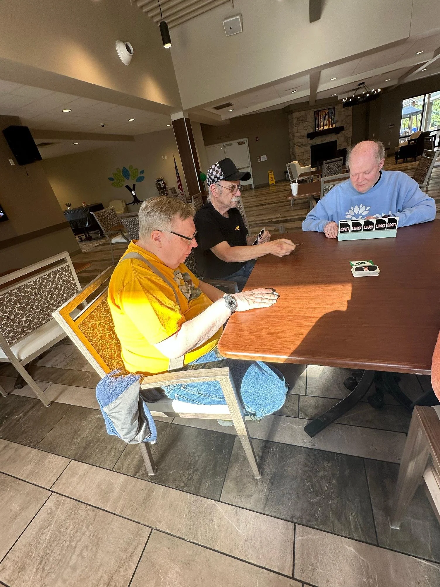 Three older men are sitting at a wooden table in a room, playing a card game. The man on the left has glasses and is wearing a yellow shirt with gray and yellow stripes. The man in the middle, with a checkered cap and glasses, is holding cards. The m
