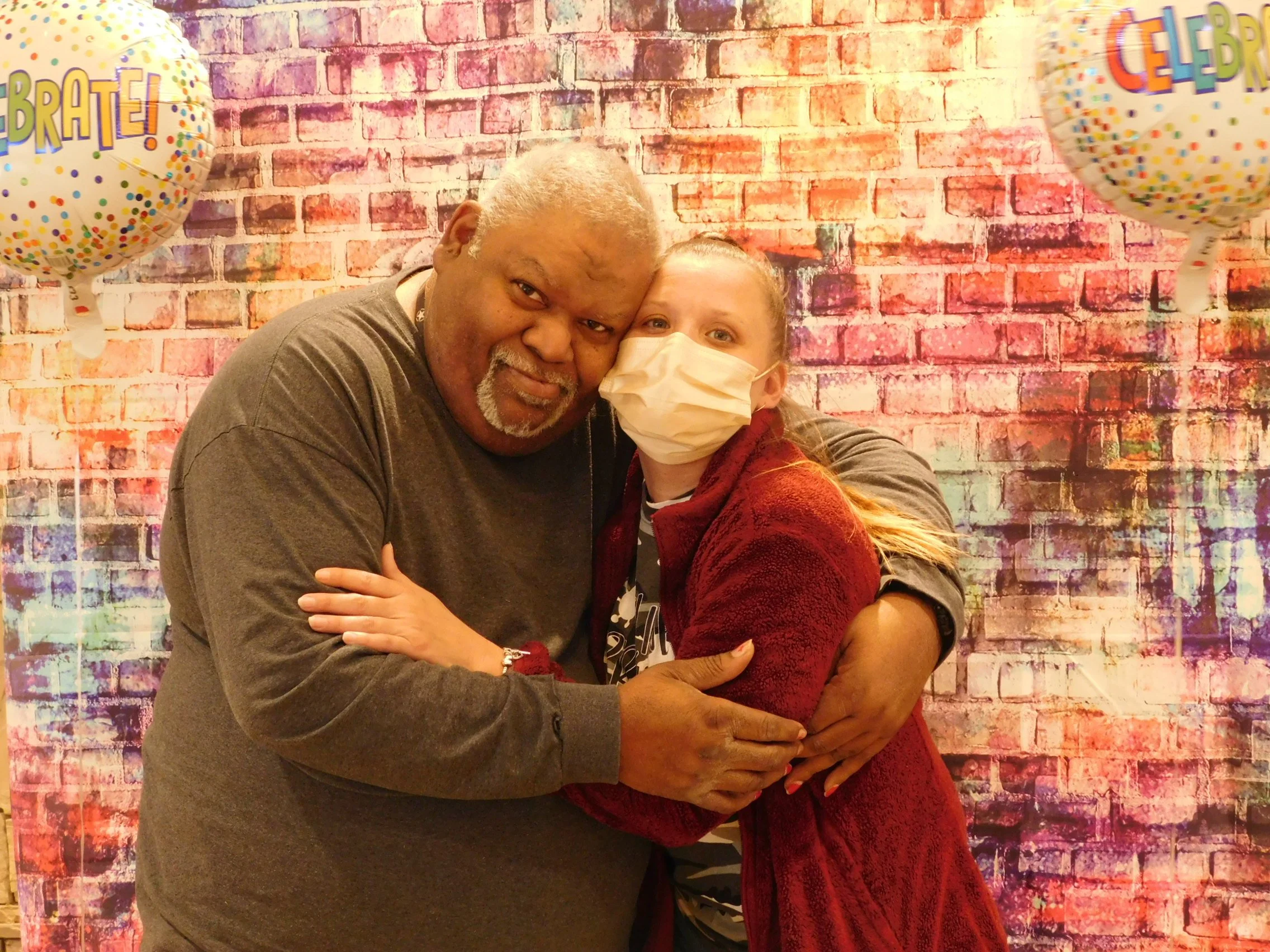 An older man with gray hair and a beard hugging a young woman with a face mask, in front of a colorful brick wall backdrop, decorated with balloons that say 'Celebrate!'