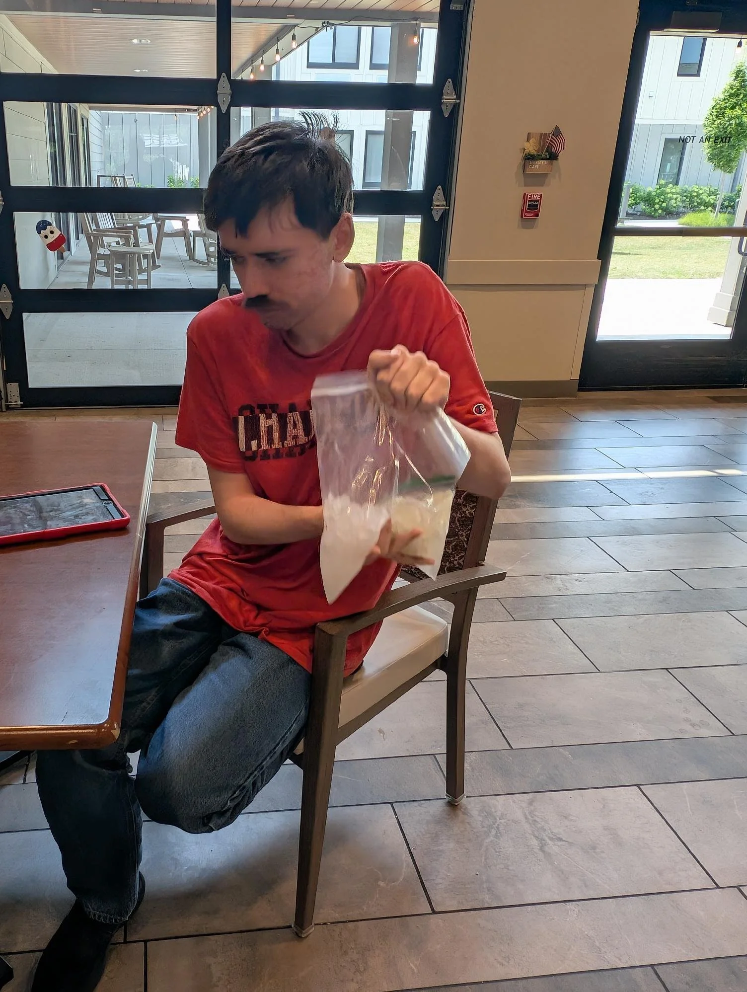 A young boy with dark hair and a red shirt sitting at a wooden table inside a restaurant. He is holding a plastic bag and looking down. The background shows glass doors and windows, with an outdoor patio area with chairs and greenery.