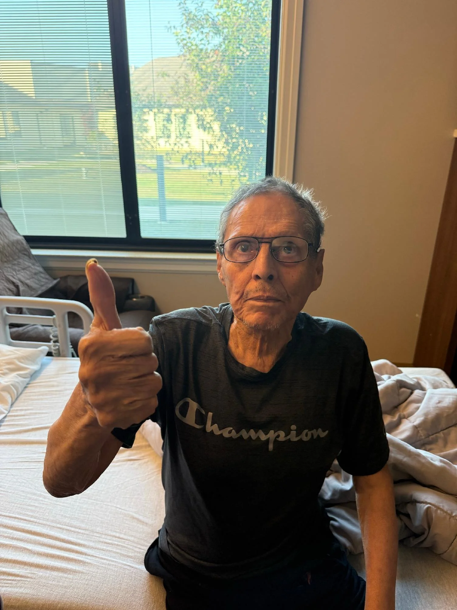 An elderly man with glasses sitting on a bed, giving a thumbs-up gesture with his right hand, wearing a black Champion t-shirt, in a room with a window in the background showing trees and houses.