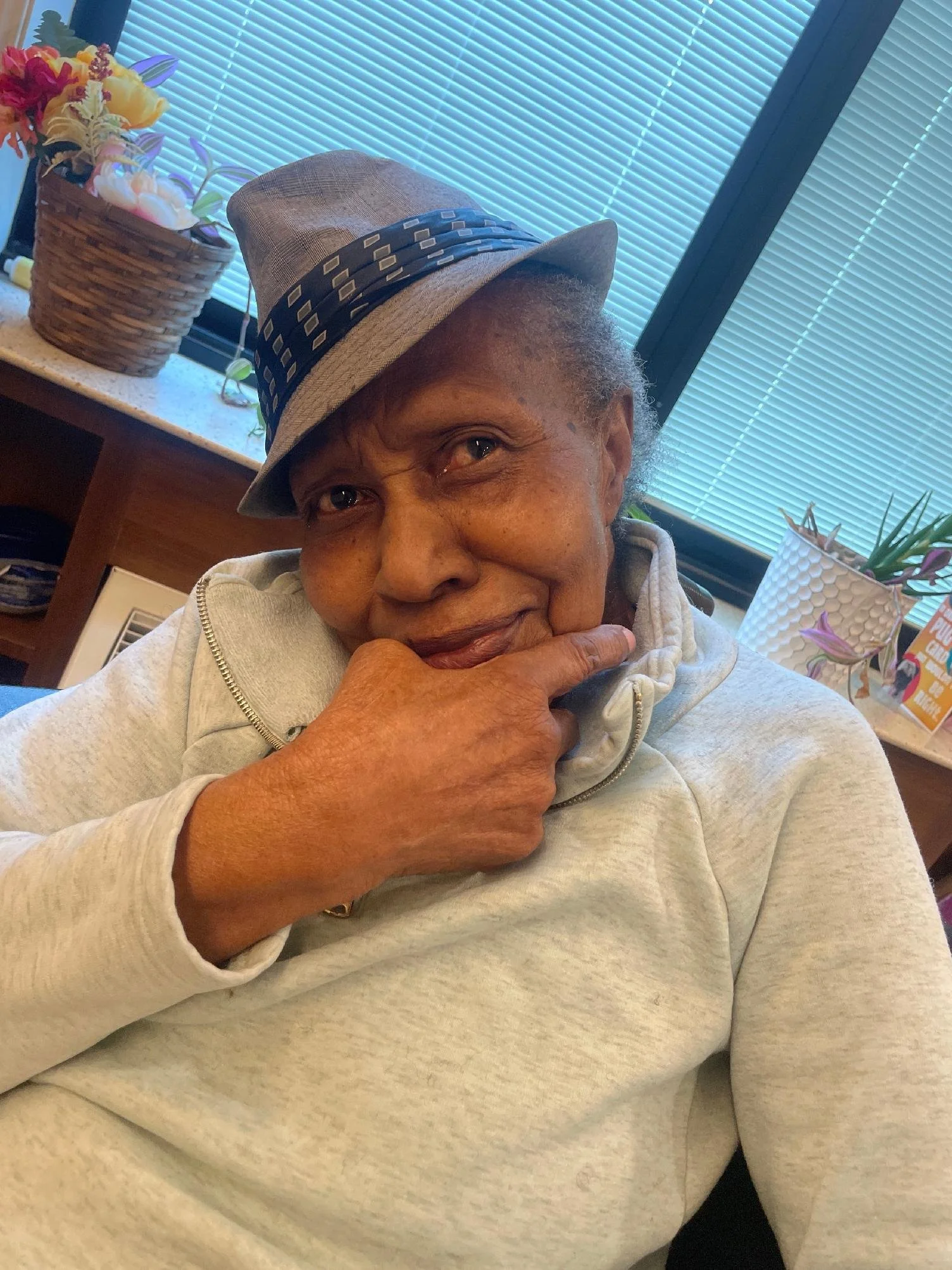 A woman wearing a gray hoodie and a fedora hat, sitting indoors near a window with closed blinds, resting her chin on her hand with a gentle smile, surrounded by potted plants and flowers.