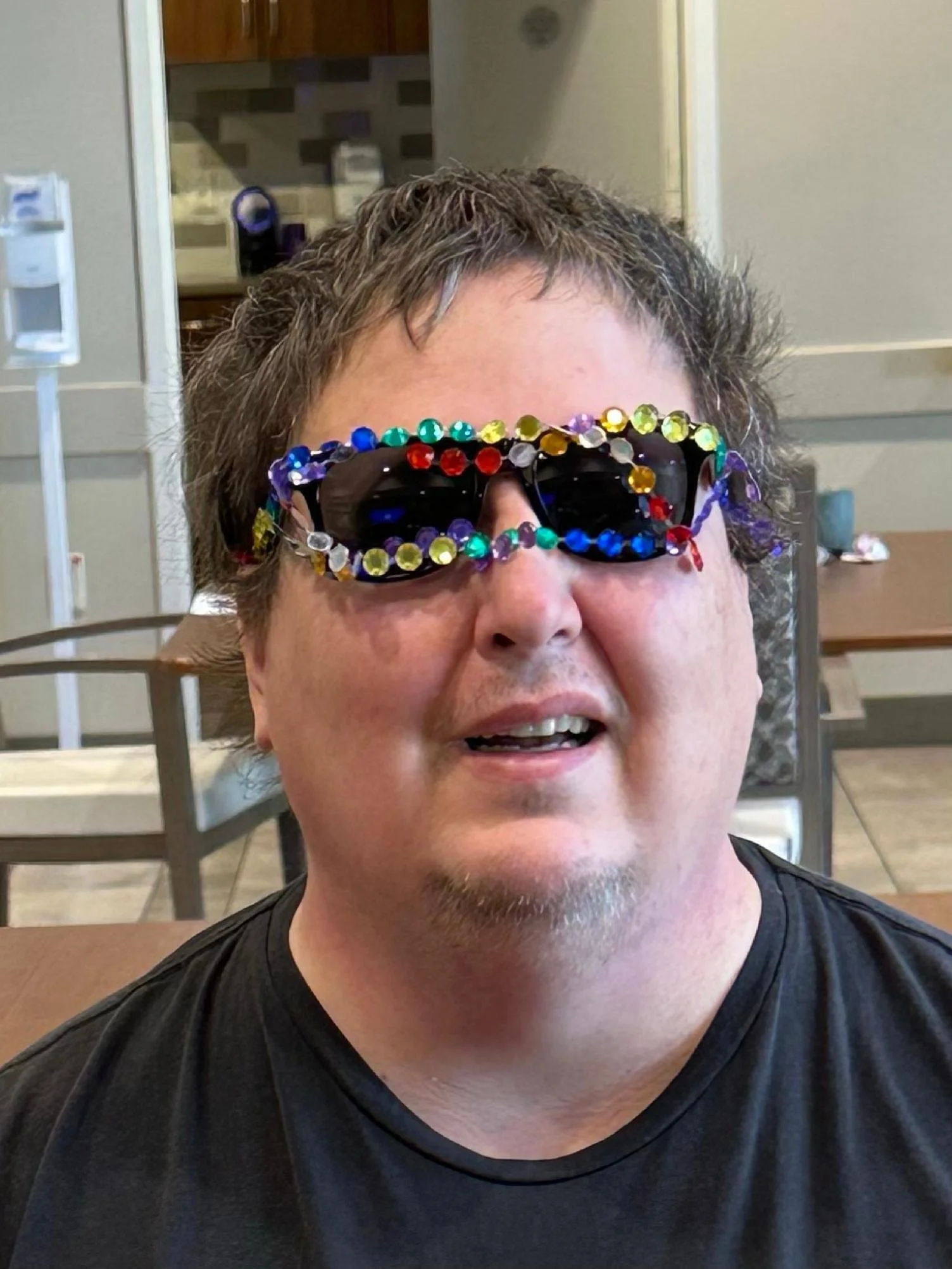 A person wearing sunglasses decorated with colorful rhinestones, sitting in a cafe or restaurant with a background that includes a kitchen area and dining furniture.