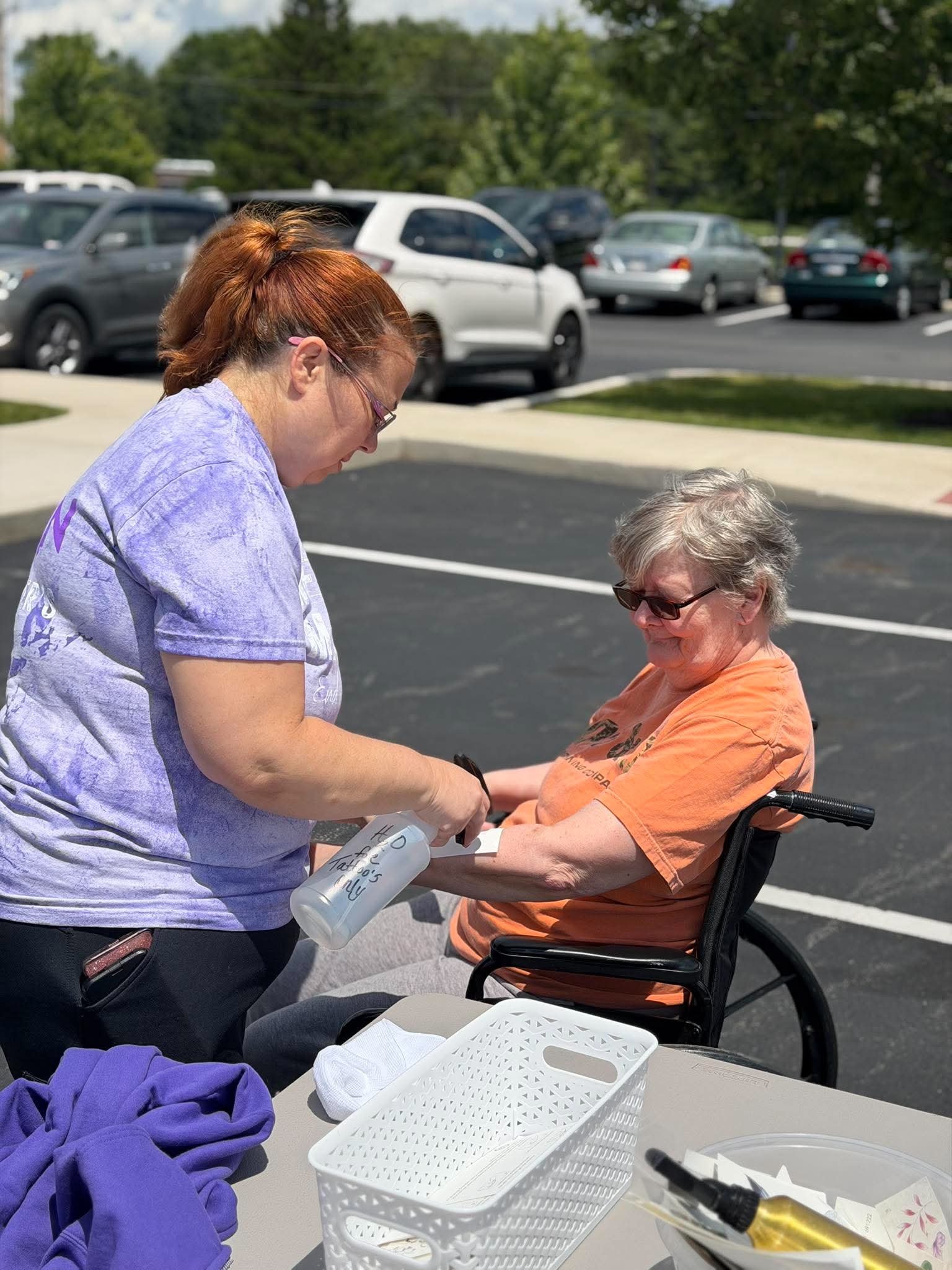 A woman in a purple shirt and glasses is standing, assisting an elderly woman with gray hair and glasses, who is sitting in a wheelchair at an outdoor table in a parking lot. The elderly woman is wearing an orange shirt and is holding a white bag. Th