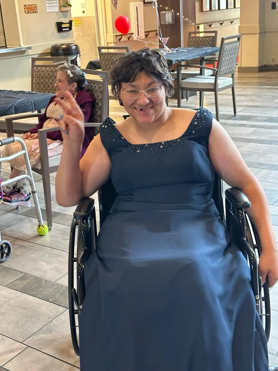 A woman with short curly hair, glasses, and a big smile sitting in a wheelchair, wearing a dark blue dress with embellishments on the shoulders. In the background, a young girl with a headband and pink dress is sitting at a table, with balloons and d