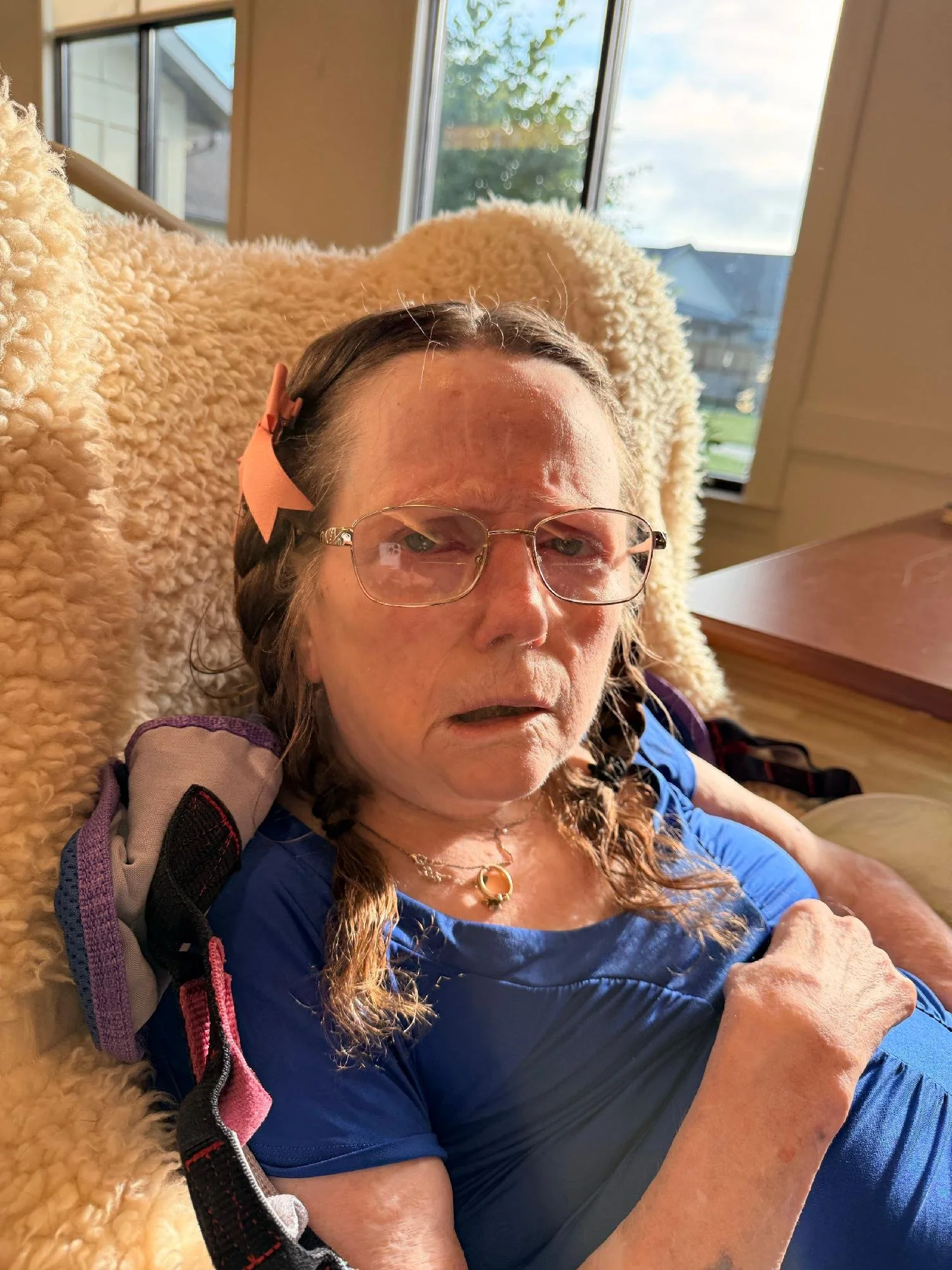 A woman with glasses, long braided hair, and a pink bow, lying on a blanket near large windows with trees visible outside, wearing a blue shirt and a necklace.