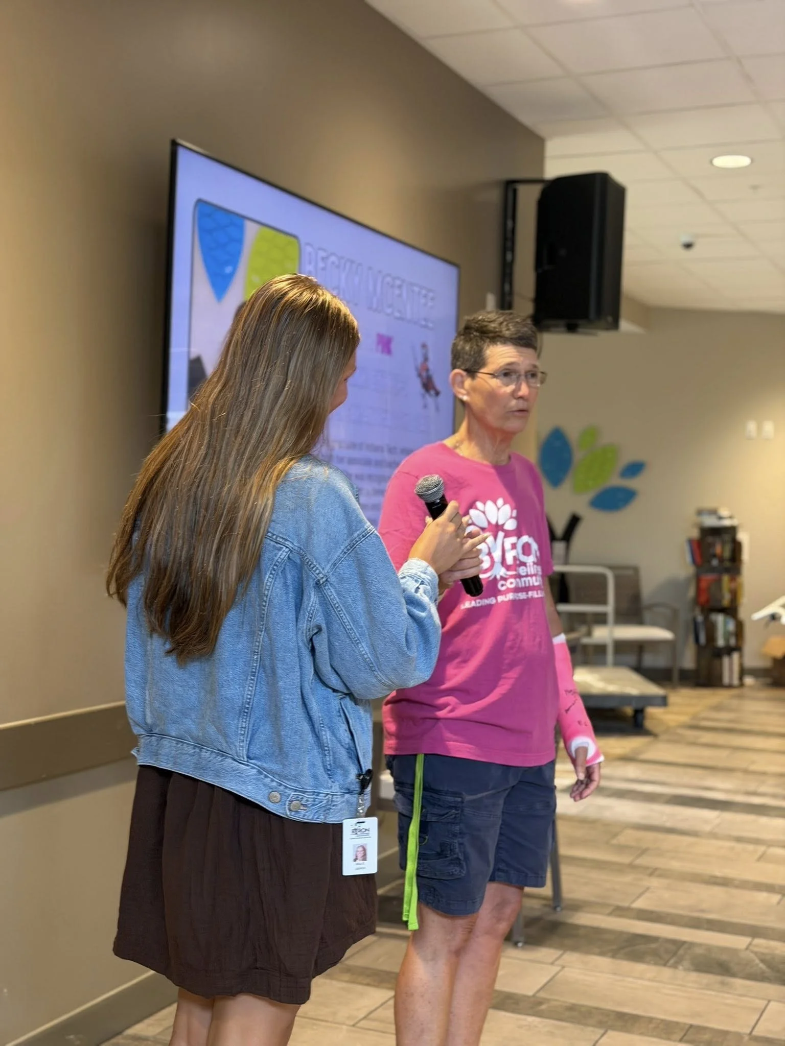 Two women standing indoors near a large screen and a speaker. One woman with long hair and a denim jacket holds a microphone, while the other woman, wearing glasses and a pink T-shirt, appears to be speaking.