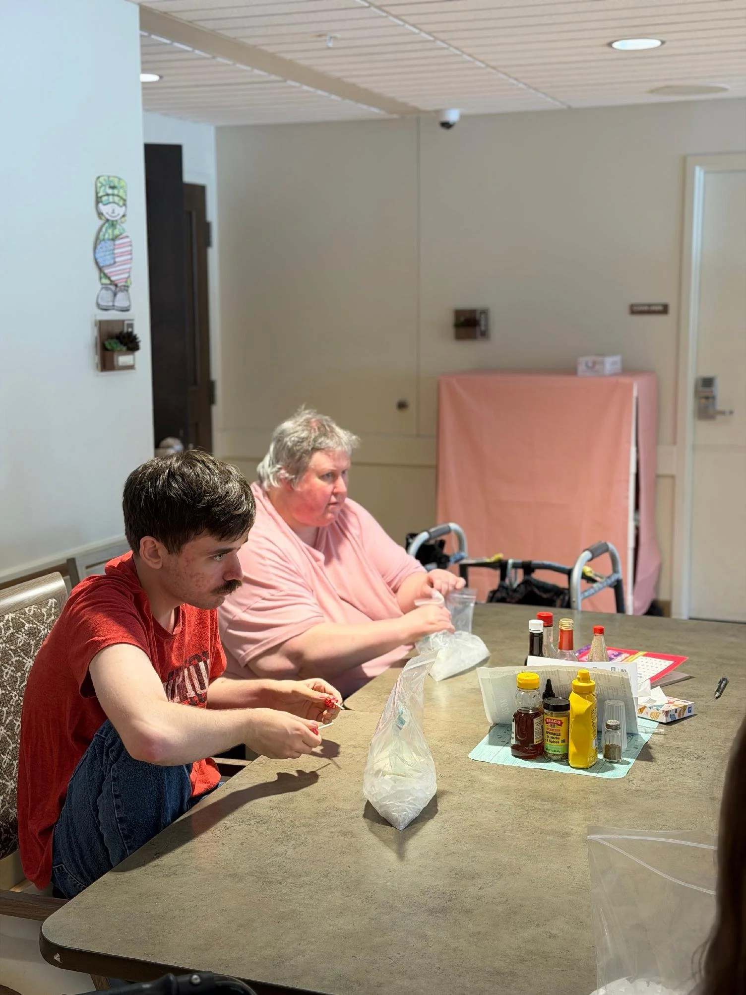 Two people sitting at a table with supplies, preparing food or craft project, in a room with beige walls and wood ceiling panels.