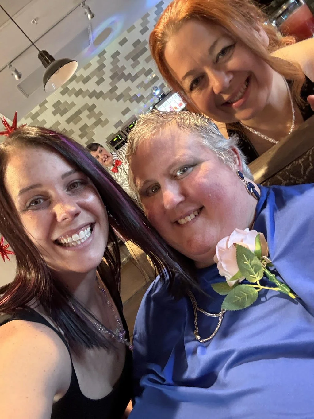 Four women smiling and posing for a photo in a restaurant or cafe, with festive decorations, including red stars on their hair, and one woman wearing a blue shirt with a purple rose pin.