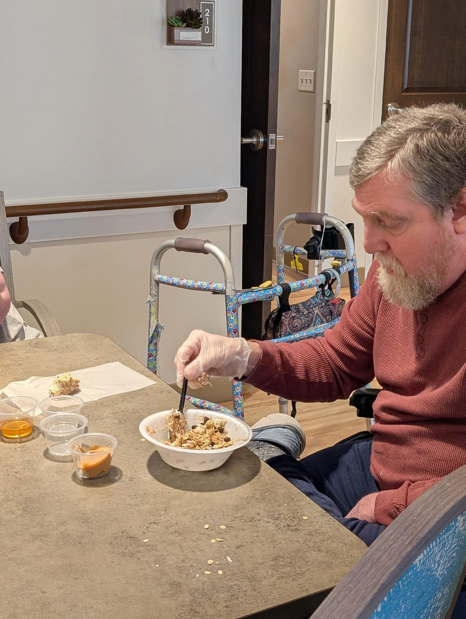 An elderly man with gray hair and a beard is sitting at a dining table, feeding himself cereal from a bowl using a spoon. There are various small containers of syrup and other toppings on the table. In the background, a walker is visible near a doorw