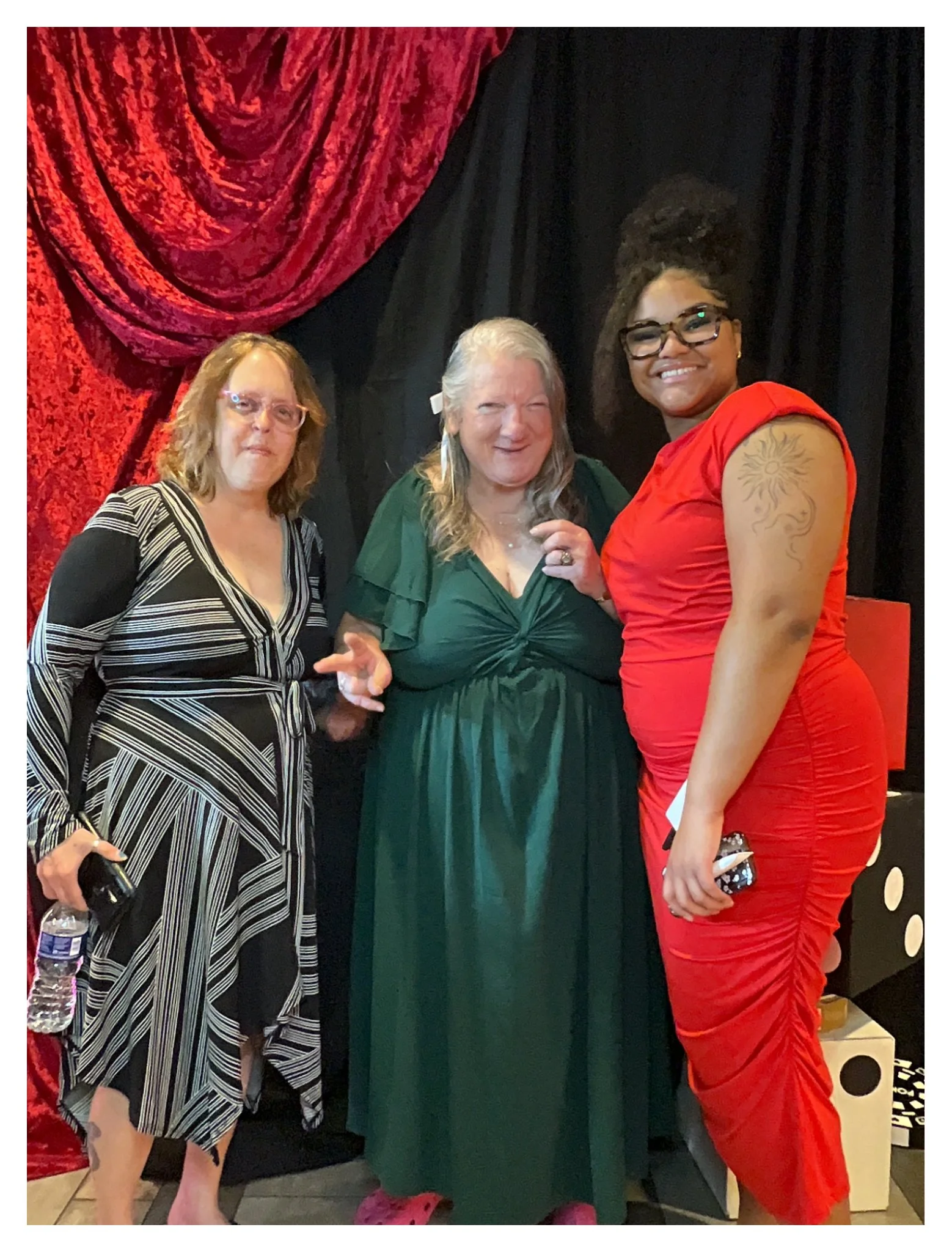 Three women standing together indoors, with a black and red curtain backdrop behind them. The woman on the left has shoulder-length red hair, wears glasses, and a black-and-white striped dress. The woman in the middle has long gray hair, wears a gree