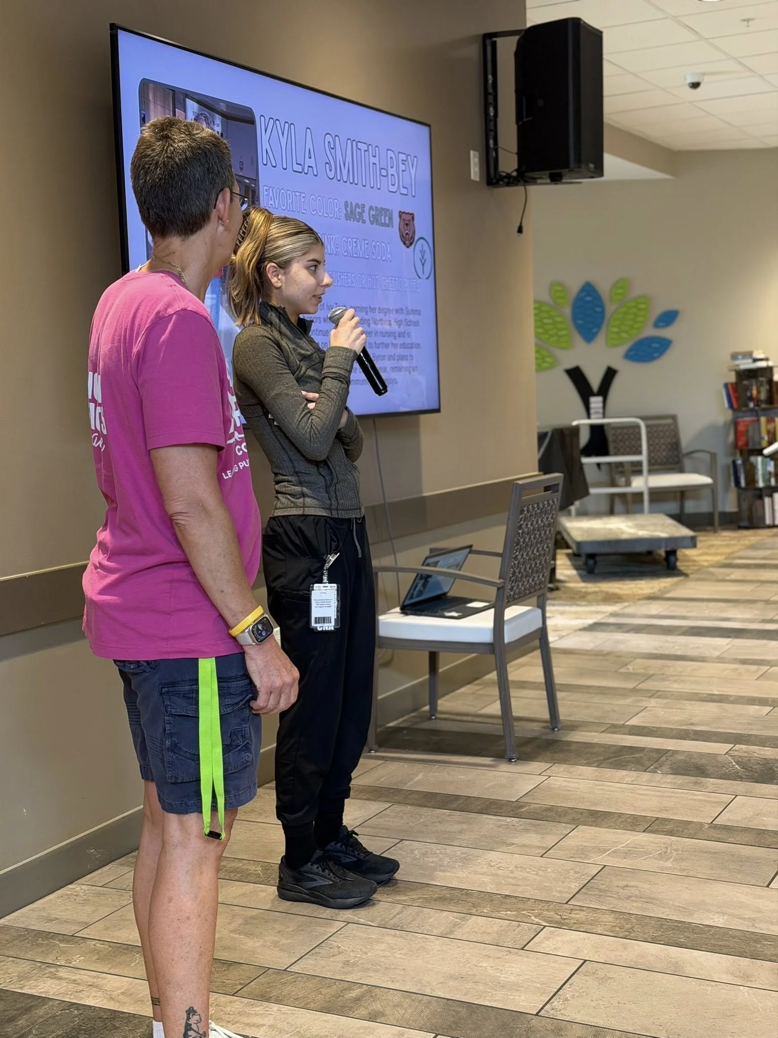 Two people standing in front of a large screen, one holding a microphone, while another looks at the screen. The screen displays information about Kyla Smith-Bey, including favorite color and drink.