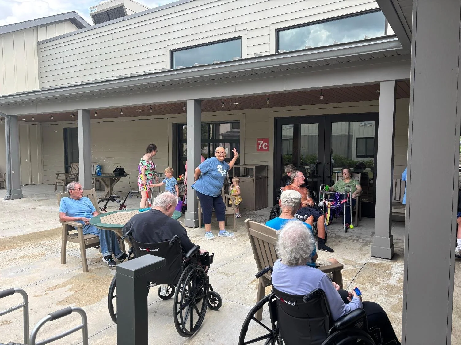 Gathering of elderly people and children outdoors in front of a building with a covered patio, some people seated in chairs and wheelchairs, and a woman standing and waving.
