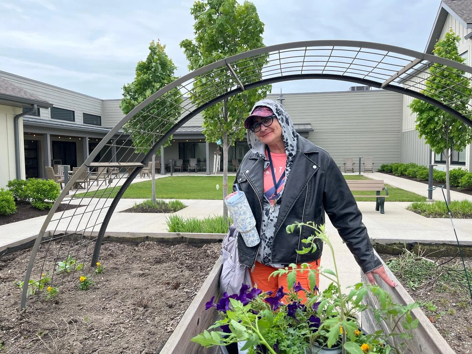 A woman with glasses wearing a black leather jacket, pink shirt, and orange pants, standing next to a garden bed with flowers and plants, in an outdoor courtyard with trees, benches, and a building with a patio.
