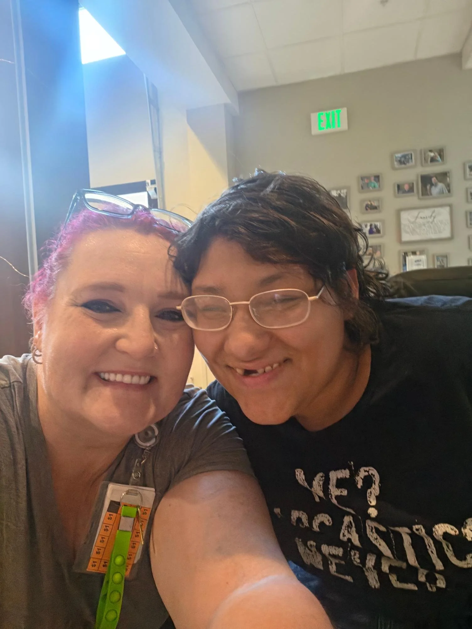 Two women smiling for a selfie in an indoor setting, one with glasses on her head and the other with glasses over her eyes, with a green exit sign and a collage of photo frames on the wall in the background.