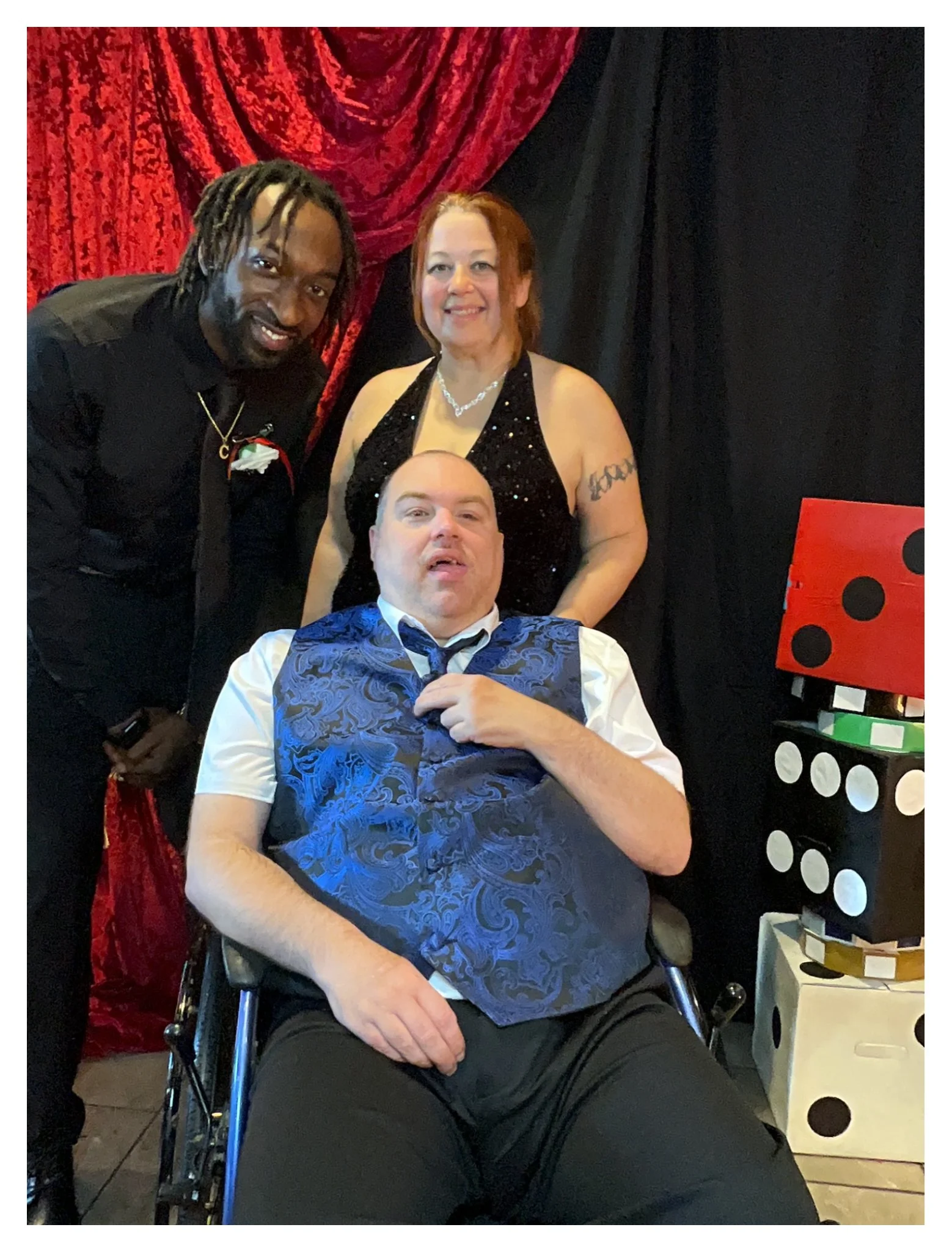 Three people dressed in formal attire, two standing and one seated in a wheelchair. The background features red and black curtains and a colorful game board.