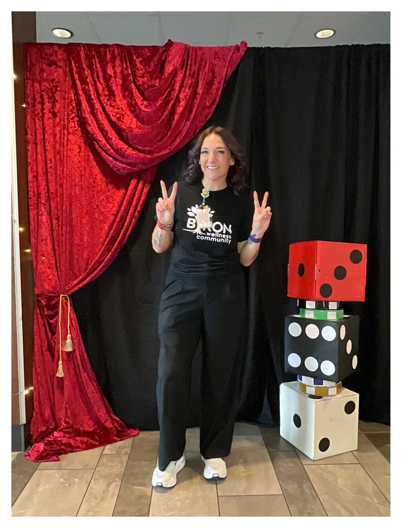 A woman standing in front of red and black curtains, making peace signs with her hands, wearing a black t-shirt, black pants, and white sneakers, with oversized dice stacked beside her.