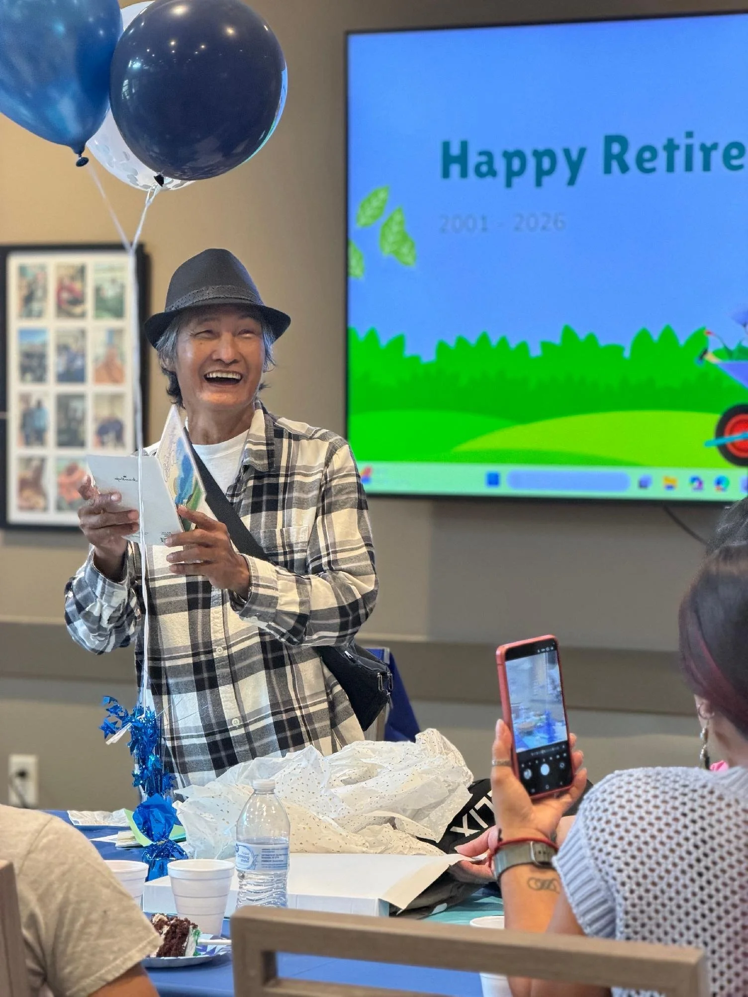 An elderly woman with gray hair, wearing a black hat and plaid shirt, is smiling while holding a card or a letter. She is at a celebration table with balloons, a water bottle, and paper decorations. A large screen in the background displays a message