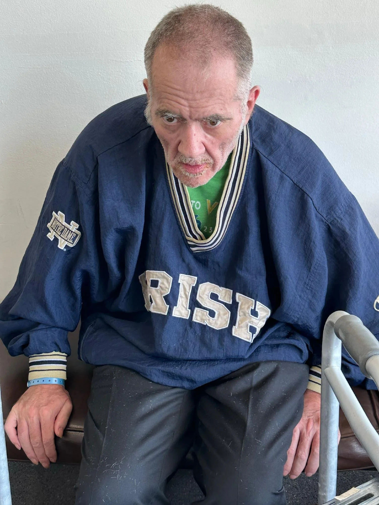 An elderly man wearing a blue hockey jersey with 'RICE' written on it, sitting and holding a crutch, with a serious expression on his face.