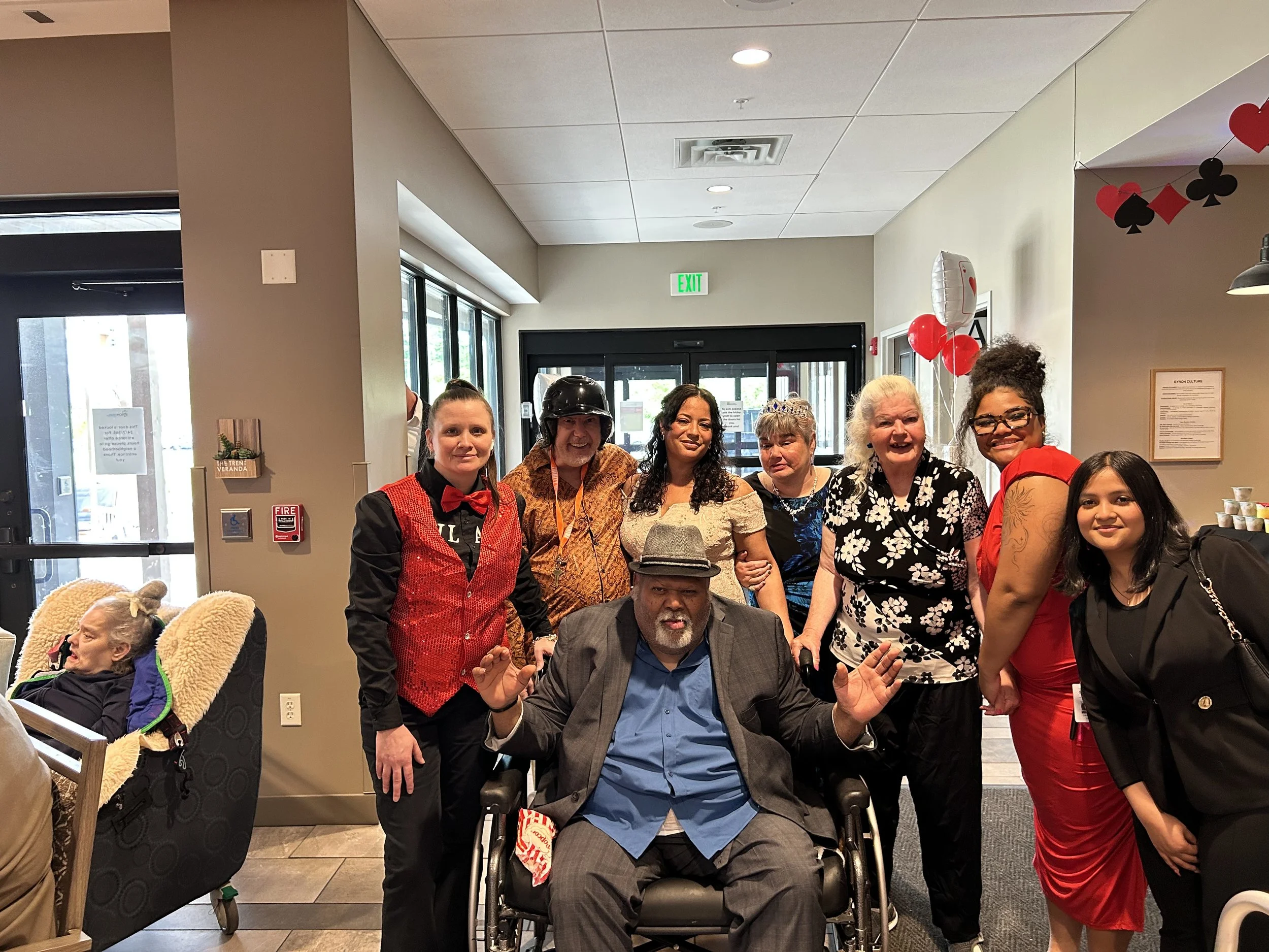 A group of people posing for a photo indoors, with some individuals standing and one person in a wheelchair sitting in the front. There are festive decorations including balloons and playing card symbols hanging from the ceiling.