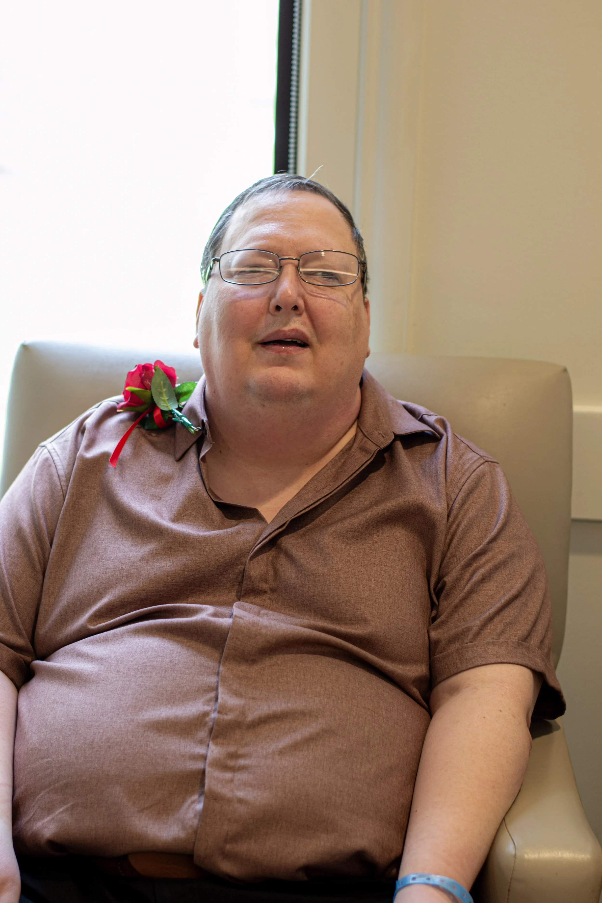 A man with glasses sitting in a beige chair, wearing a brown shirt, with a red flower on his left shoulder and a blue wristband on his right wrist, in front of a window with bright daylight.