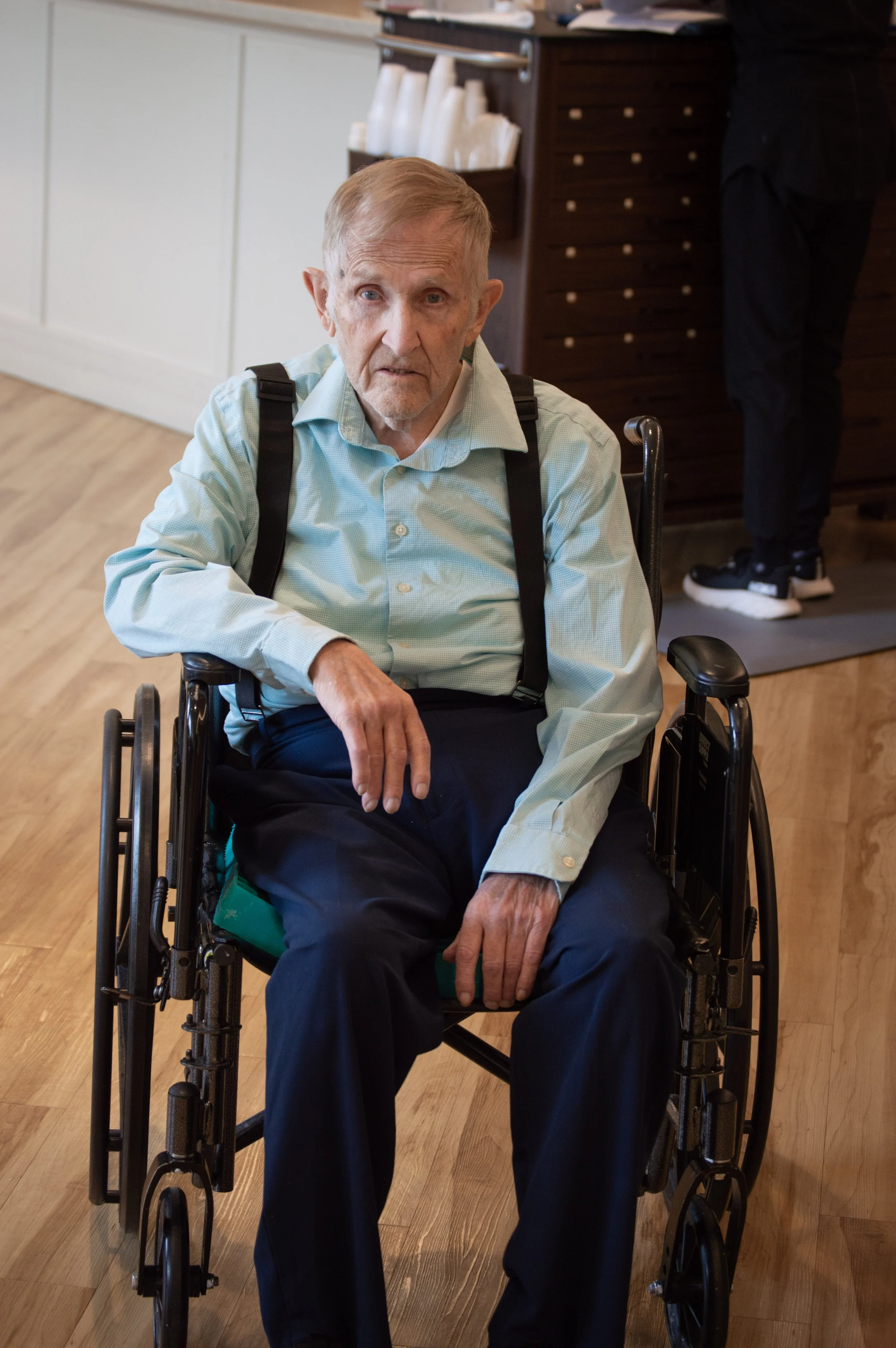 An elderly man sitting in a wheelchair inside a room, wearing a light blue shirt and dark pants, with a woman in black standing in the background near a wooden counter.