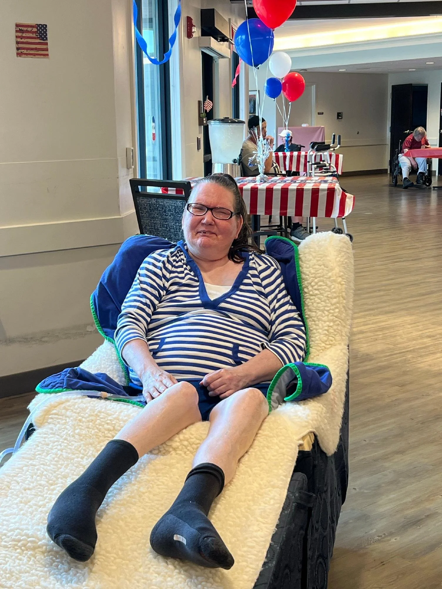 A woman in a wheelchair at a celebration with red, white, and blue balloons and striped tablecloths.
