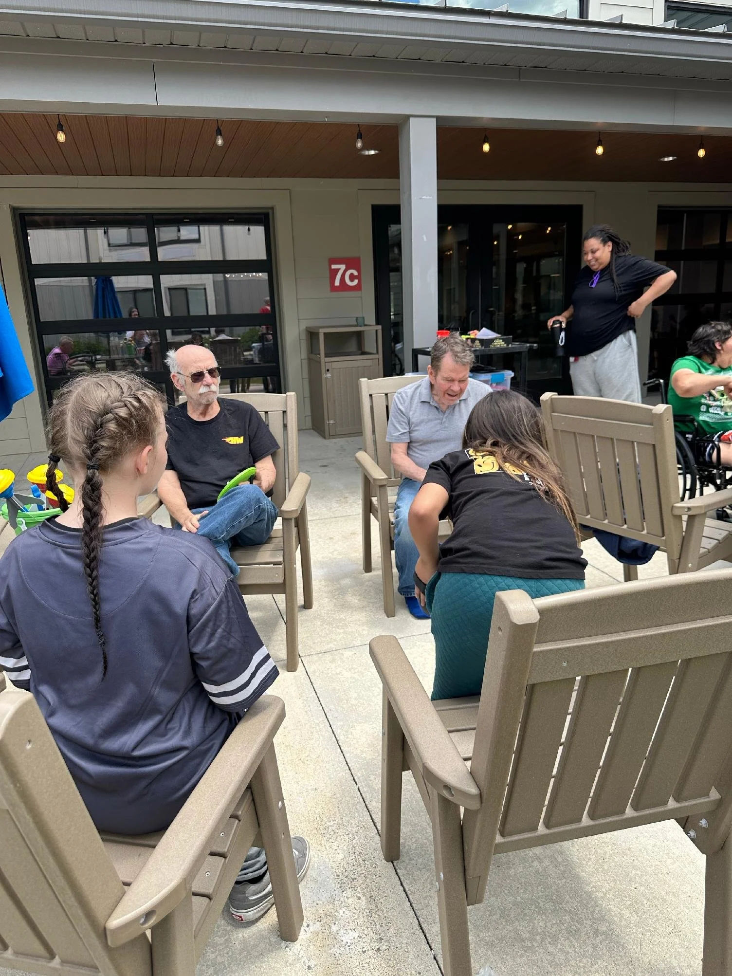 Group of people sitting on outdoor patio furniture, engaging in conversation, with some standing and others seated. A building with large windows and a patio area is in the background.