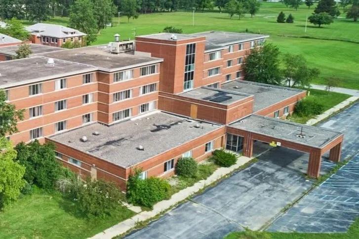Aerial view of a four-story brick apartment complex with a parking lot and surrounding green lawn with trees.